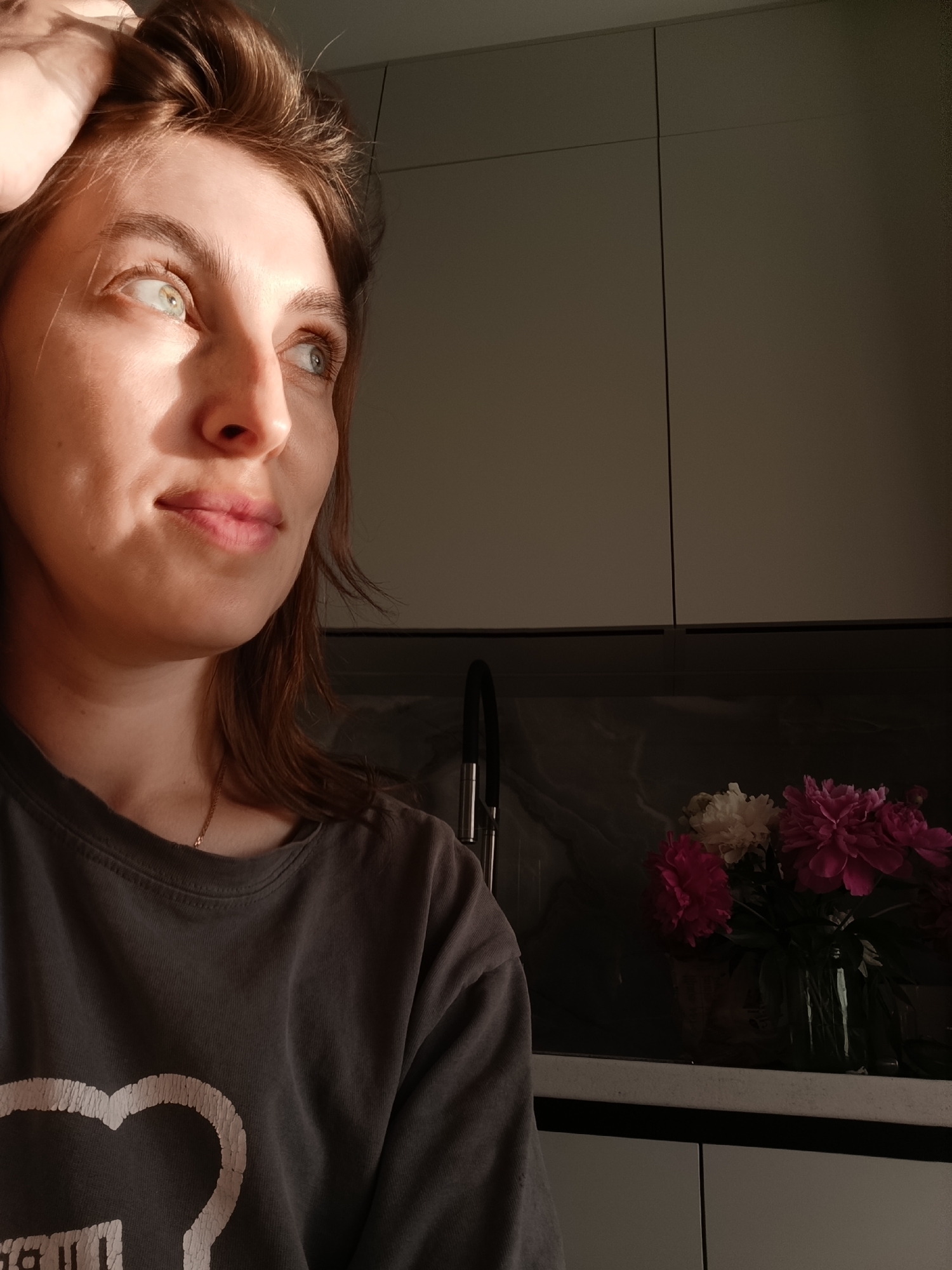 A medium close-up shot captures a woman with brown hair touching her head with her left hand. She is looking off to her right, with sunlight highlighting the left side of her face. She is wearing a dark gray t-shirt with a white graphic design on it. In the background, to the right of the woman, is a kitchen counter with a vase of pink and white flowers. Above the counter are cabinets. The lighting suggests it is either early morning or late afternoon. This scene is located in the city of Chaikovsky, Russia.