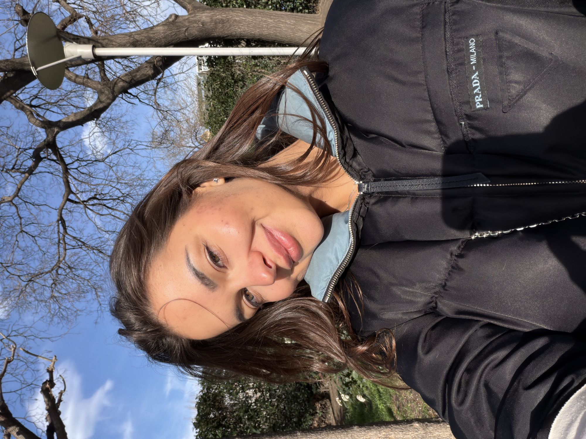 A young woman poses for a selfie in Tbilisi, Georgia. She wears a black Prada puffer jacket with a light blue inner lining. Trees and a lamppost with a round light fixture are visible in the background. The sky is blue with some clouds.