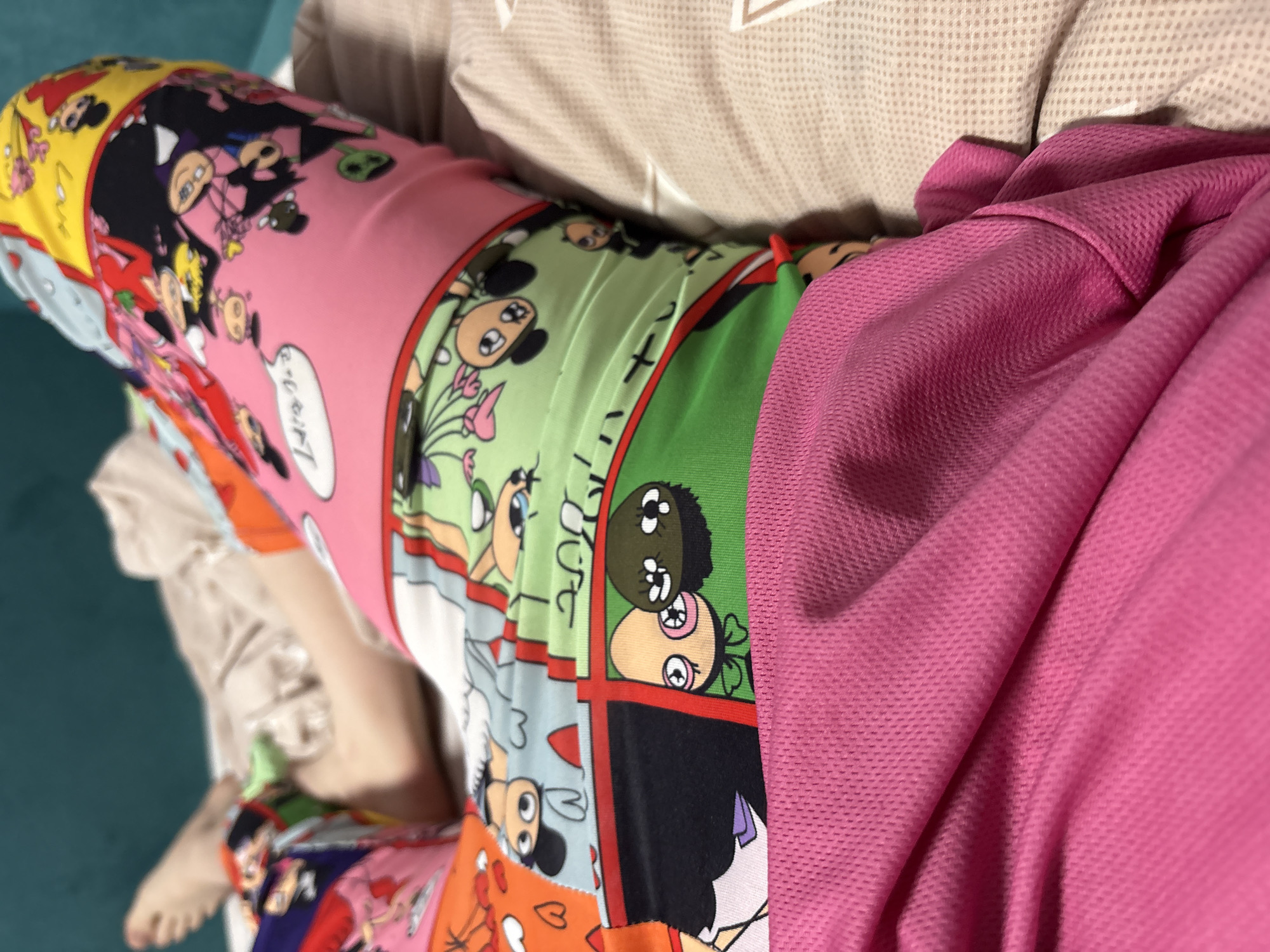 The image shows a person lying on a bed. The person is wearing colorful, cartoon-themed pajamas. A pink shirt or hoodie is draped over part of the pajamas and the person's body. The bed has a light-colored patterned pillow or headboard. The location is Прикамский, Russia, cannot be determined from the image.