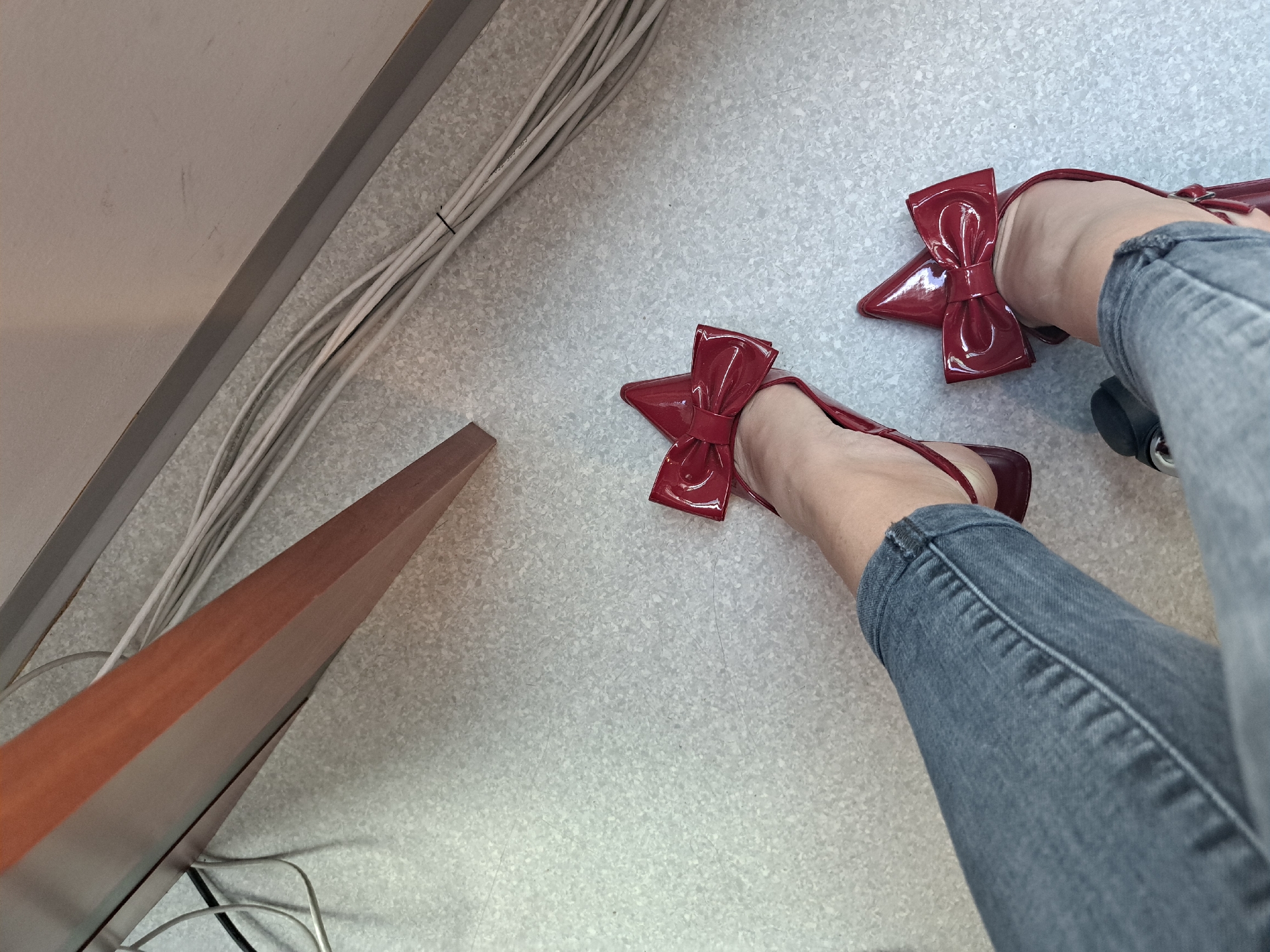 The image shows a pair of feet wearing red patent leather shoes with bow details, positioned on a light grey linoleum floor. The person is wearing grey jeans. In the periphery, there are visible wires, a desk or cabinet panel in wood, and a section of wall. No location in Sofia, Bulgaria, can be determined from the image.