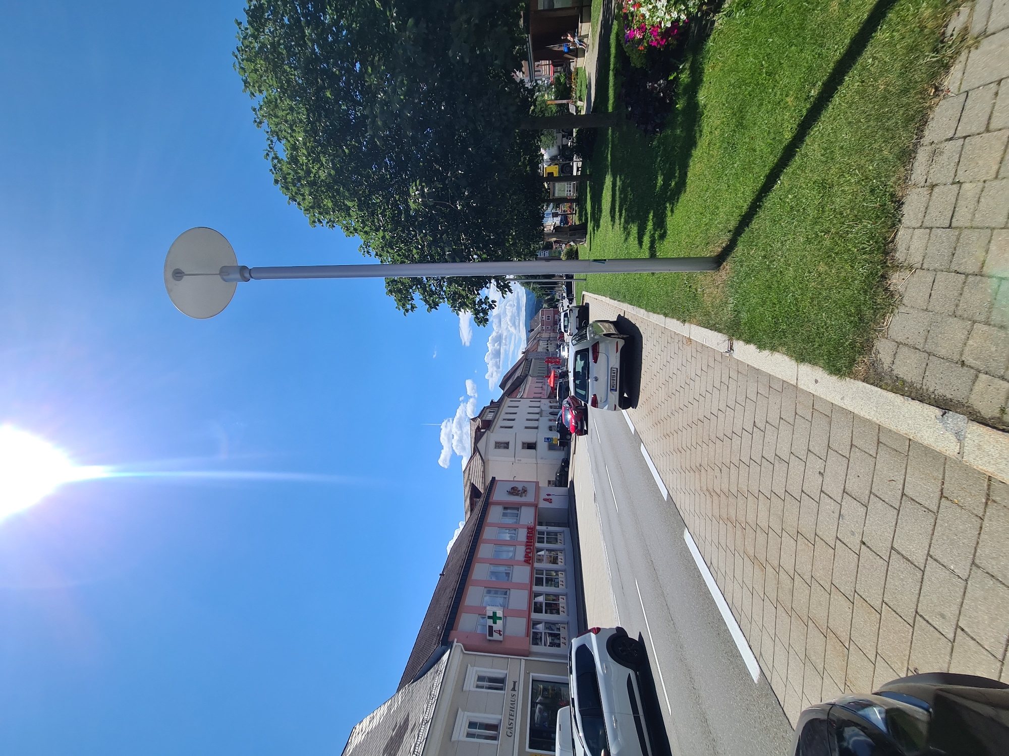 The image depicts a street scene in Sankt Marein bei Neumarkt, Austria, on a bright, sunny day with a clear blue sky and a few white clouds. The perspective is from a low angle, looking down a street lined with buildings on the left and a grassy area with trees and a lamppost on the right.
On the left side of the street, there are several buildings. The closest building on the left has a sign that reads "GÄSTEHAUS" (Guesthouse). Next to it is a building with a pink and white facade, featuring a green cross symbol and "APOTHEKE" (Pharmacy) written on it. Further down the street, more buildings are visible, with varied architectural styles and colors. Cars are parked along the street and some are driving. A white SUV is parked on the left, and several other cars, including a white Volkswagen Golf, are on the road and parked on the right side of the street.
The right side of the scene features a well-maintained green lawn with mature trees casting shadows. A modern lamppost stands prominently in the grassy area. In the background of the grassy area, there are some wooden structures, possibly benches or shelters, and some people are visible sitting or walking. A variety of flowers, including pink and red ones, are planted near the edge of the grassy area. The street itself has a paved sidewalk with interlocking stones, and a road with clear lane markings. The overall impression is of a clean and orderly urban environment.