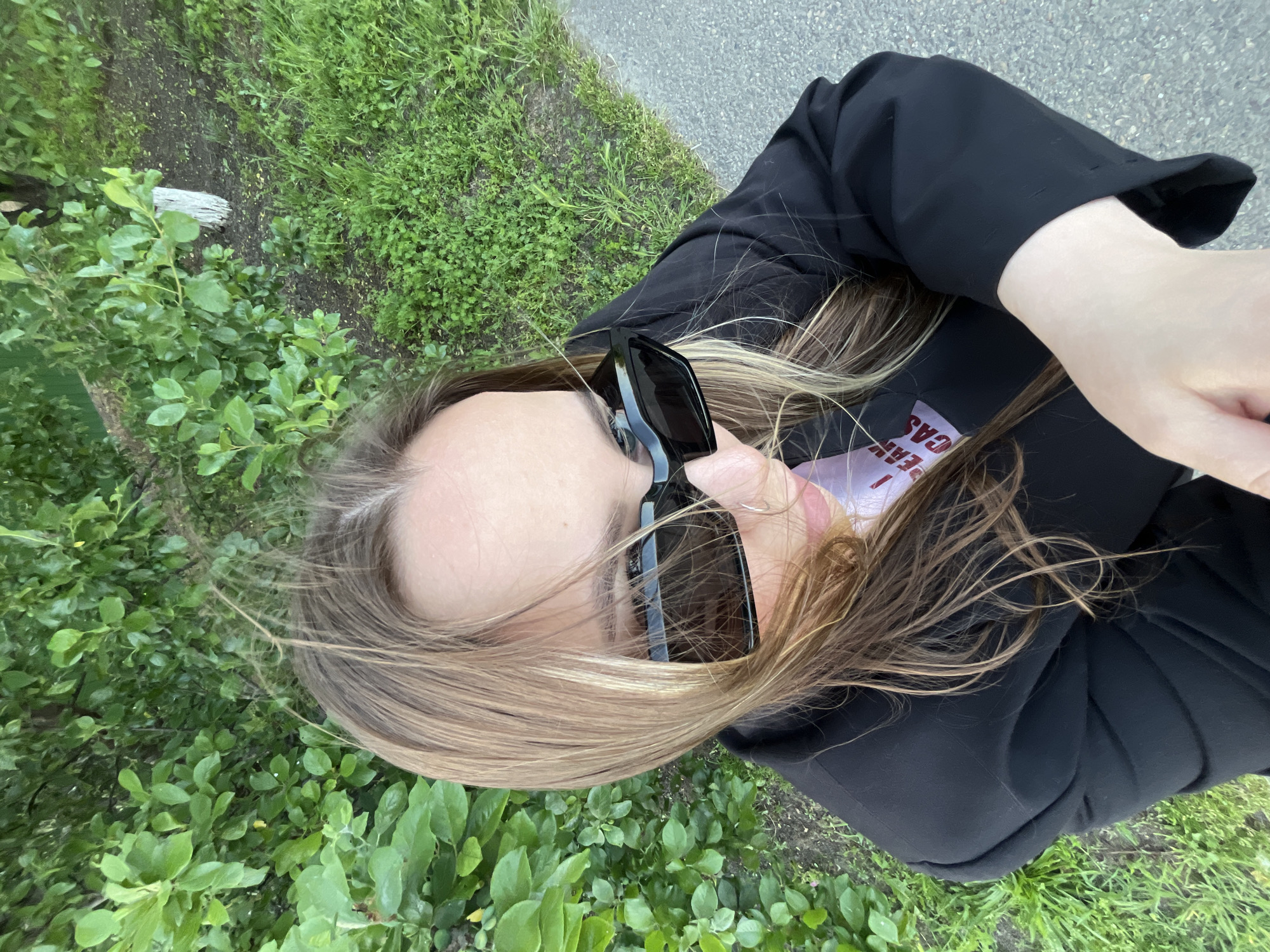 A woman with brown hair is taking a selfie outdoors. She is wearing sunglasses and a black jacket. The background consists of grass and pavement. The location is not discernible from the image.