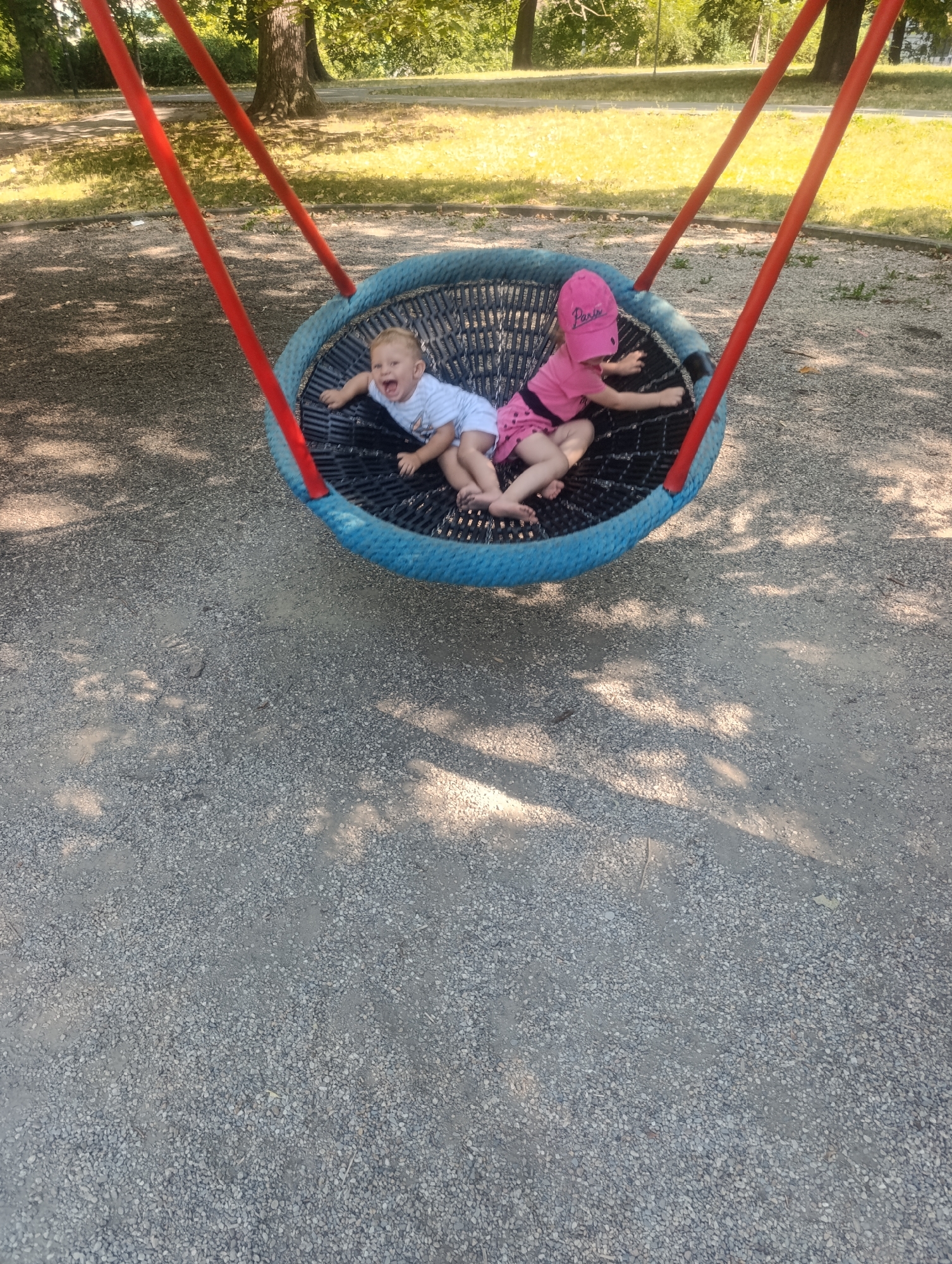 Two children are sitting on a blue, circular swing with red supporting poles in a park. The child on the left is a young boy with blond hair wearing a light blue and white striped t-shirt and shorts. He is lying on his back with his mouth open and eyes wide. The child on the right is a young girl wearing a pink hat and a pink dress. She is lying on her side, facing away from the camera. The swing is suspended over a gravel surface, and dappled sunlight filters through the trees in the background. The location appears to be a playground in a park.