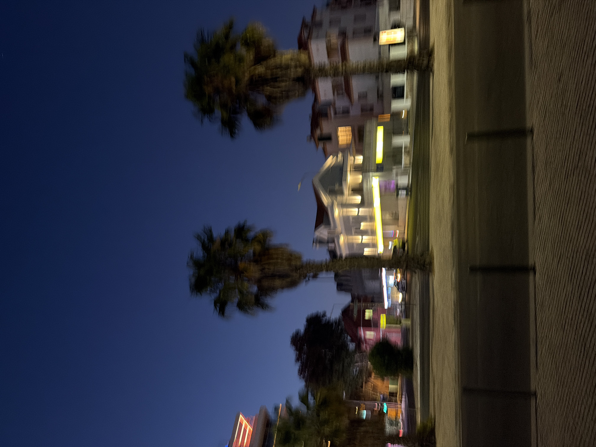 The image depicts a street scene in Cascais, Portugal at dusk or night. Tall palm trees are a prominent feature, with their fronds blurred due to motion. Buildings line the street, some illuminated with interior and exterior lights, suggesting businesses or residences. A modern building with red and white lights is visible on the left, while more traditional structures with red roofs and illuminated windows are seen further down the street. Cars are present, though indistinct due to the blur. The sky is a deep blue, indicating the late hour. The foreground shows a paved walkway with distinct linear patterns, separated from the street by dark bollards. The overall impression is a vibrant urban environment at night, captured with a slight motion blur.