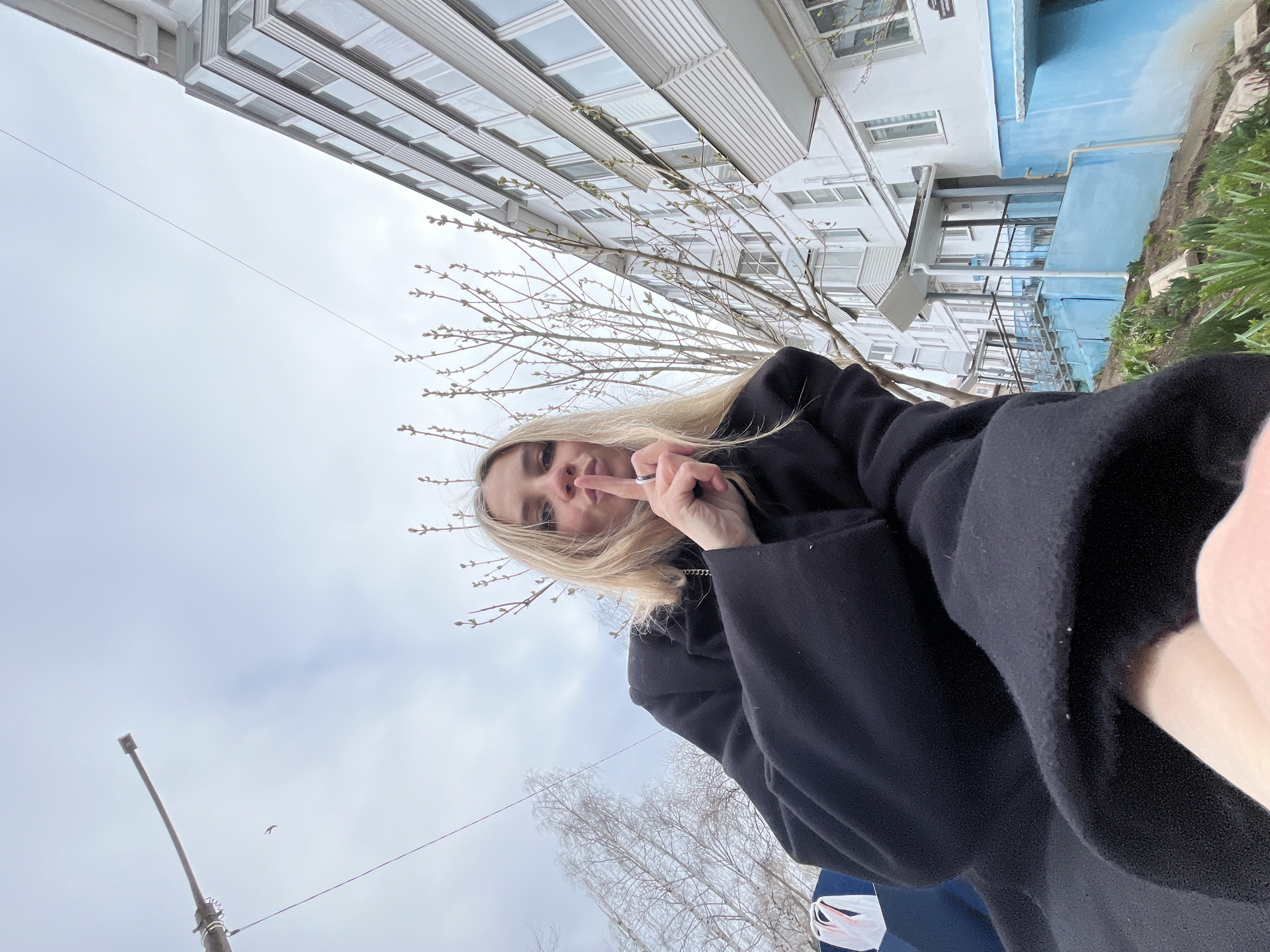 This tilted, low-angle image captures a young woman and an urban landscape under an overcast sky in Chaikovsky, Russia.
The primary subject is a young woman with long blonde hair, seen from the chest up, looking directly at the camera. She is wearing a dark, heavy coat or jacket. Her left hand (from the viewer's perspective) is raised, with her index and middle fingers extended in a "V" or peace sign. A ring is visible on her finger. Her expression appears neutral or slightly playful.
The background features a tall, light-colored apartment building with multiple windows, which appears on the left when the image is mentally oriented upright. A section of the building to the right has a vibrant blue-painted facade. Bare tree branches with small, emerging buds are scattered across the scene, indicating early spring. Some green foliage, possibly grass or bushes, is visible near the base of the blue building section.
The sky is uniformly overcast, suggesting daytime with diffuse light. A thin wire, likely a utility line, stretches across the sky. On the lower left, a dark street light pole is visible, and a small bird is captured in flight near it. A white plastic bag with red details is partially visible at the bottom right. No discernible text is present in the image.