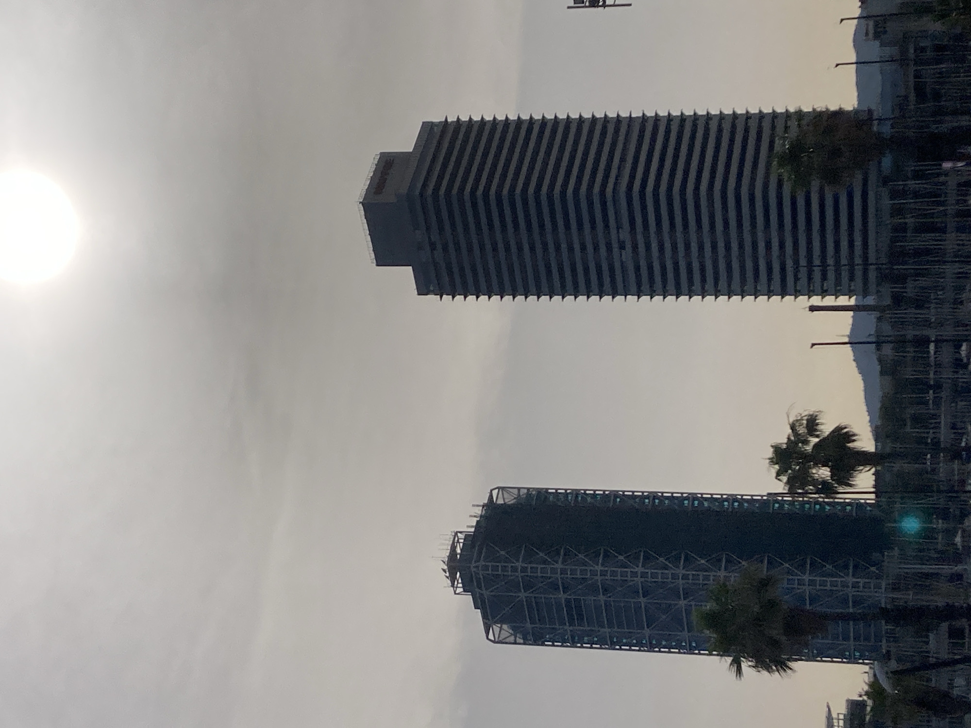 The image depicts a view of Barcelona's skyline, focusing on two prominent skyscrapers. The left skyscraper is under construction, wrapped in scaffolding and dark netting. The right skyscraper, the Hotel Arts, is a dark, horizontal-banded building with a distinct flat top. The sun is high in the hazy sky, creating a bright, diffused light. In the foreground, palm trees are visible, and the lower part of the image shows a marina with numerous masts of sailboats. In the distance, hills are faintly visible under the overcast sky. The scene appears to be captured during daylight hours, likely a hazy or partly cloudy day.