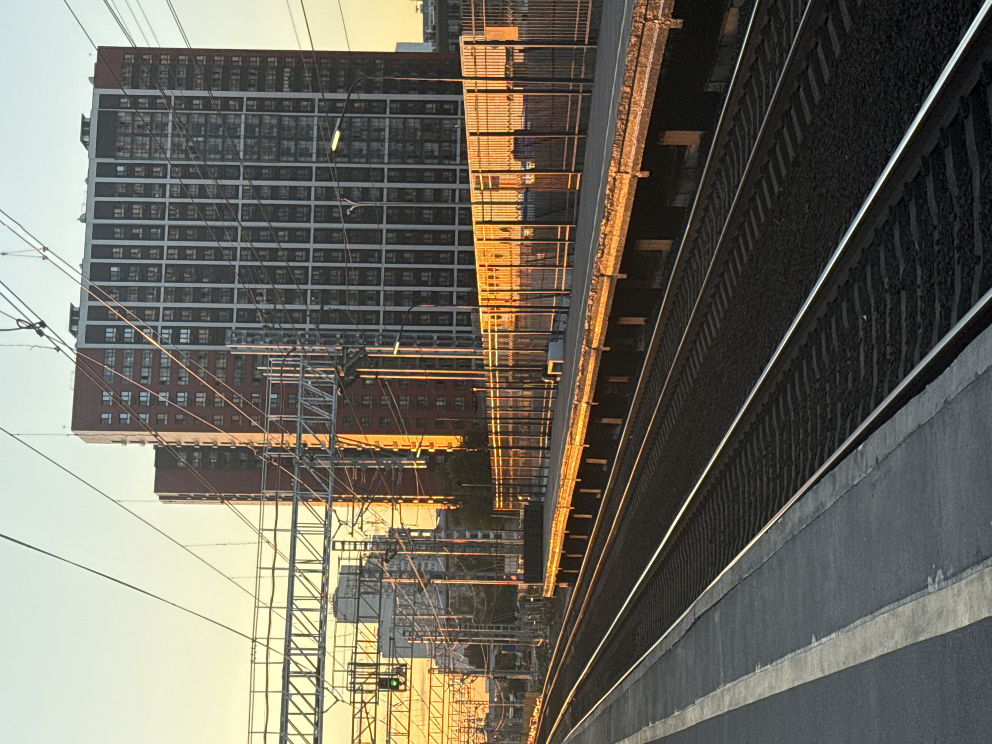 The image depicts an urban scene with a focus on a railway and a tall residential building. The perspective is from a platform next to train tracks, which are prominent in the foreground, running diagonally across the frame. The tracks are made of dark ballast and feature two parallel metal rails. Adjacent to the tracks is the platform, constructed from concrete with a distinct gray color and a white line marking the edge.
Behind the platform and tracks, a large, multi-story residential building dominates the background. Its facade is characterized by a grid-like pattern of numerous windows. The building appears to be made of brick and concrete. Another, shorter building is visible in front of the larger one, also featuring many windows.
A complex network of overhead power lines and support structures for the railway infrastructure is visible, stretching across the scene. The lighting suggests it is either early morning or late afternoon, with the golden hue of the setting or rising sun casting long shadows and illuminating the scene. The sky is clear and a pale yellow or orange, indicative of warm, low sunlight.
The overall mood is one of urban infrastructure and daily life, with the setting suggesting a train station in Khimki, Russia, given the context. There are no visible people, and the activity appears to be the quiet moment before or after a train's passage, or simply the ambient state of the location. The image captures the interplay of architectural elements and the functional aspects of a transportation hub under specific lighting conditions.