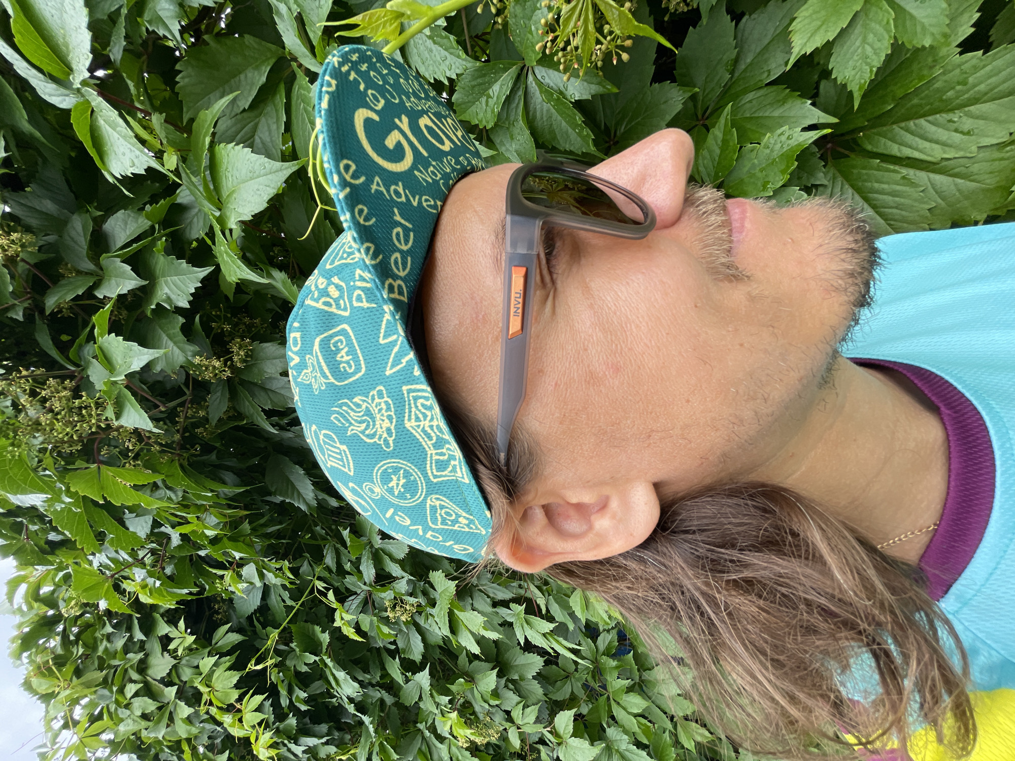 The image shows a close-up, profile view of a person from the nose up, standing in front of a lush green leafy vine. The person is wearing sunglasses with gray frames and orange accents on the side that read "INVU." On their head, they are wearing a teal baseball cap with a yellow pattern that includes images of pizza, a compass, a campfire, and the words "Beer" and "Gravel." The person has long, brown hair and a mustache and goatee. The background is dominated by the dense green foliage of the vine, with some small, light-colored berries visible. The lighting suggests it may be overcast or late in the day. The overall setting appears to be outdoors.