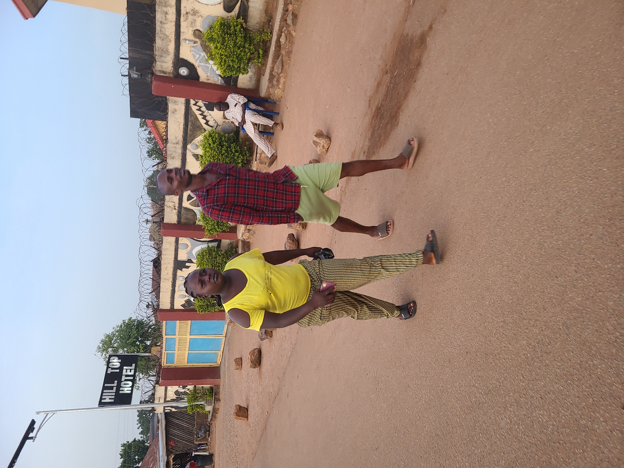 Two men stand on a paved road in Jos, Nigeria. One wears a yellow t-shirt and yellow-striped pants, holding a pink object, and the other wears a red plaid shirt and green shorts. Both wear sandals. Behind them is a painted wall topped with barbed wire, and a sign reads "HILL TOP HOTEL." A man sits on a blue chair near the wall. Bushes are planted near the wall, and the sky is visible.