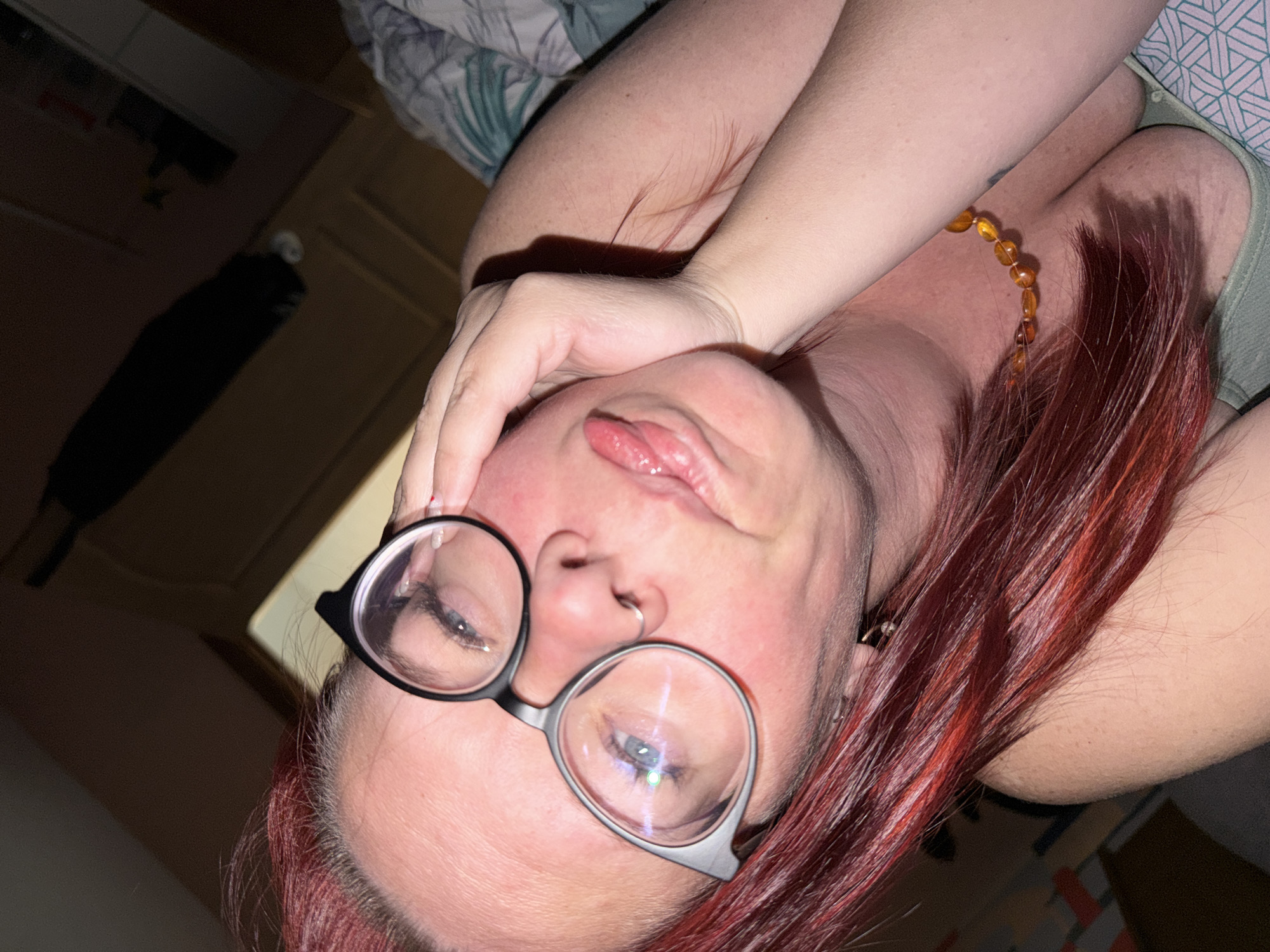A woman with red hair, wearing eyeglasses with black frames, is reclining. She is wearing a necklace of amber-colored beads. Her tongue is slightly extended. The setting appears to be indoors, near a bed. The location is not discernible from the provided image.