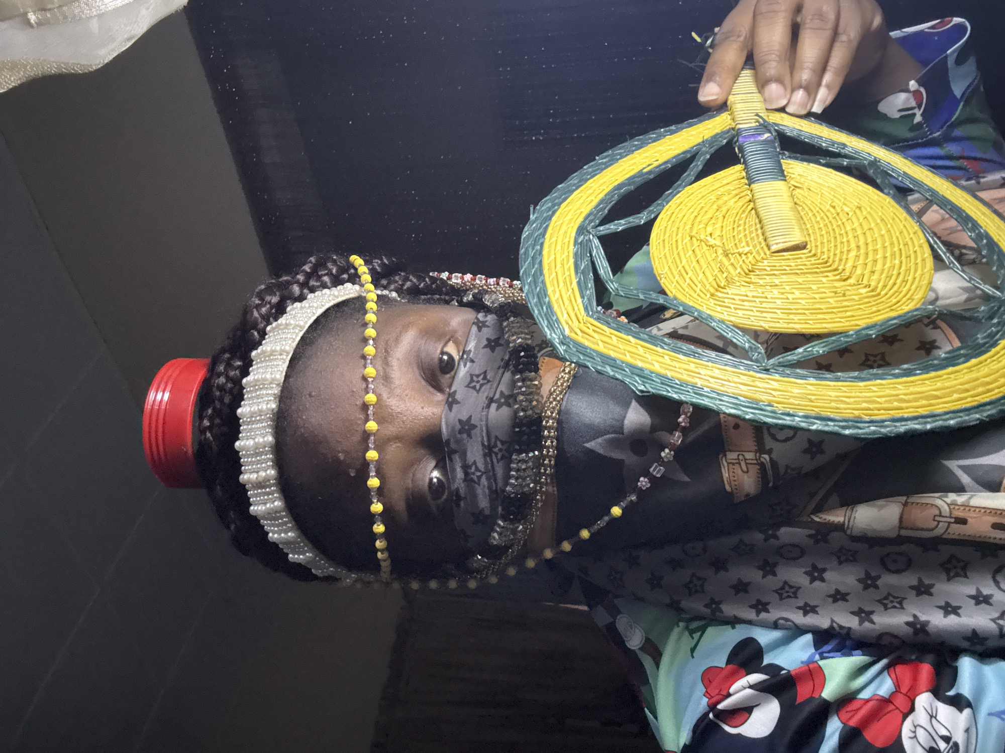 A woman is holding a yellow and green woven fan. She is wearing a pearl headband, yellow beaded necklace, and a gray face covering. Her clothing includes a gray patterned shirt, and a lower layer of blue fabric with Disney characters. The location is likely a room in Madangala, Nigeria.