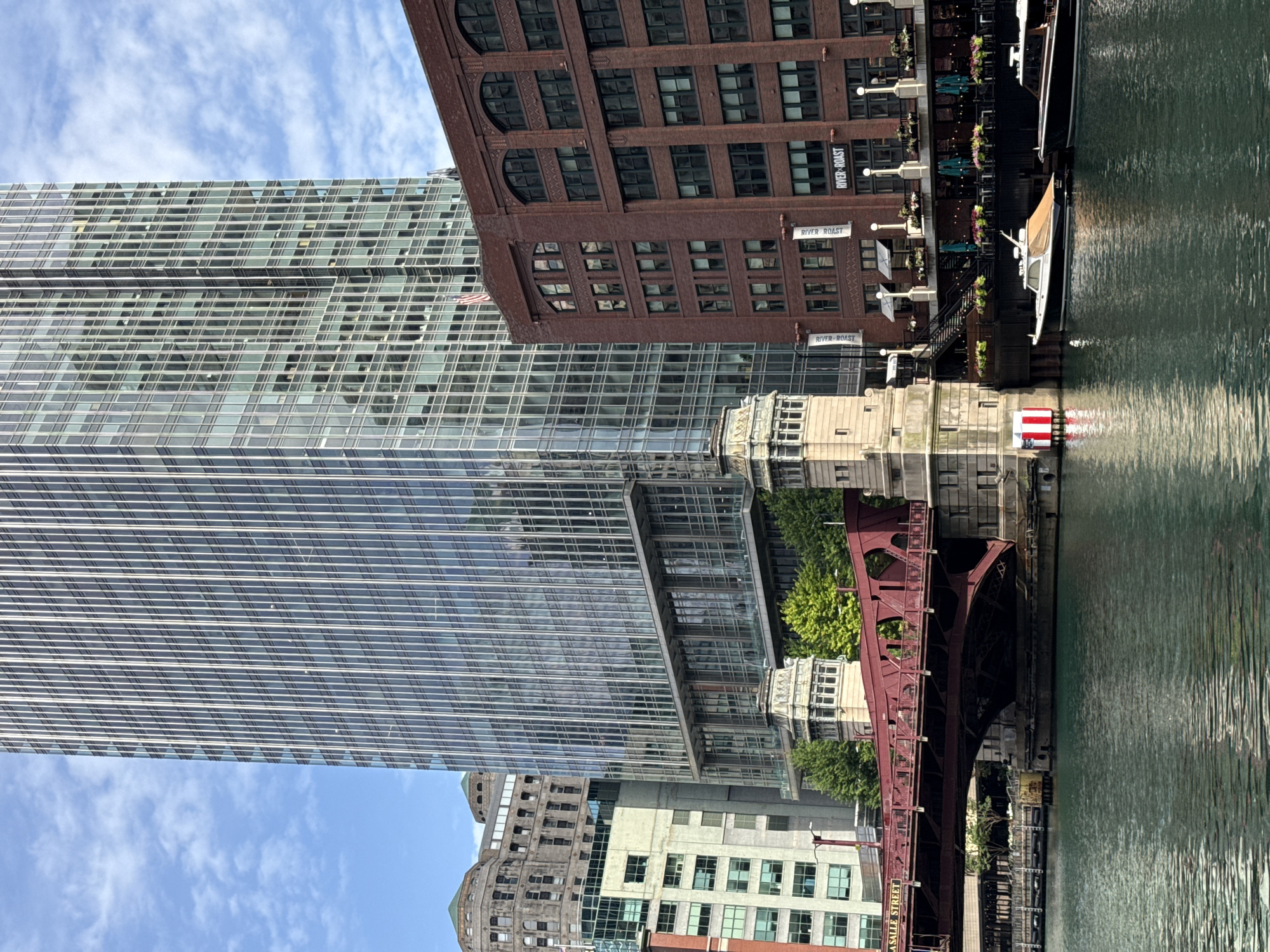 The image shows a view of the Chicago River in Chicago, Illinois, USA, on a cloudy day. A red truss bridge, identified by a sign as the "LaSalle Street" bridge, spans the river. To the right of the bridge are several modern and older buildings. The most prominent is a tall, glass-facade skyscraper. Adjacent to it is a red brick building with arched windows. In the foreground, two boats are docked along the riverbank. The river itself is a deep green color, with gentle ripples on its surface. The sky above is blue with scattered white clouds.