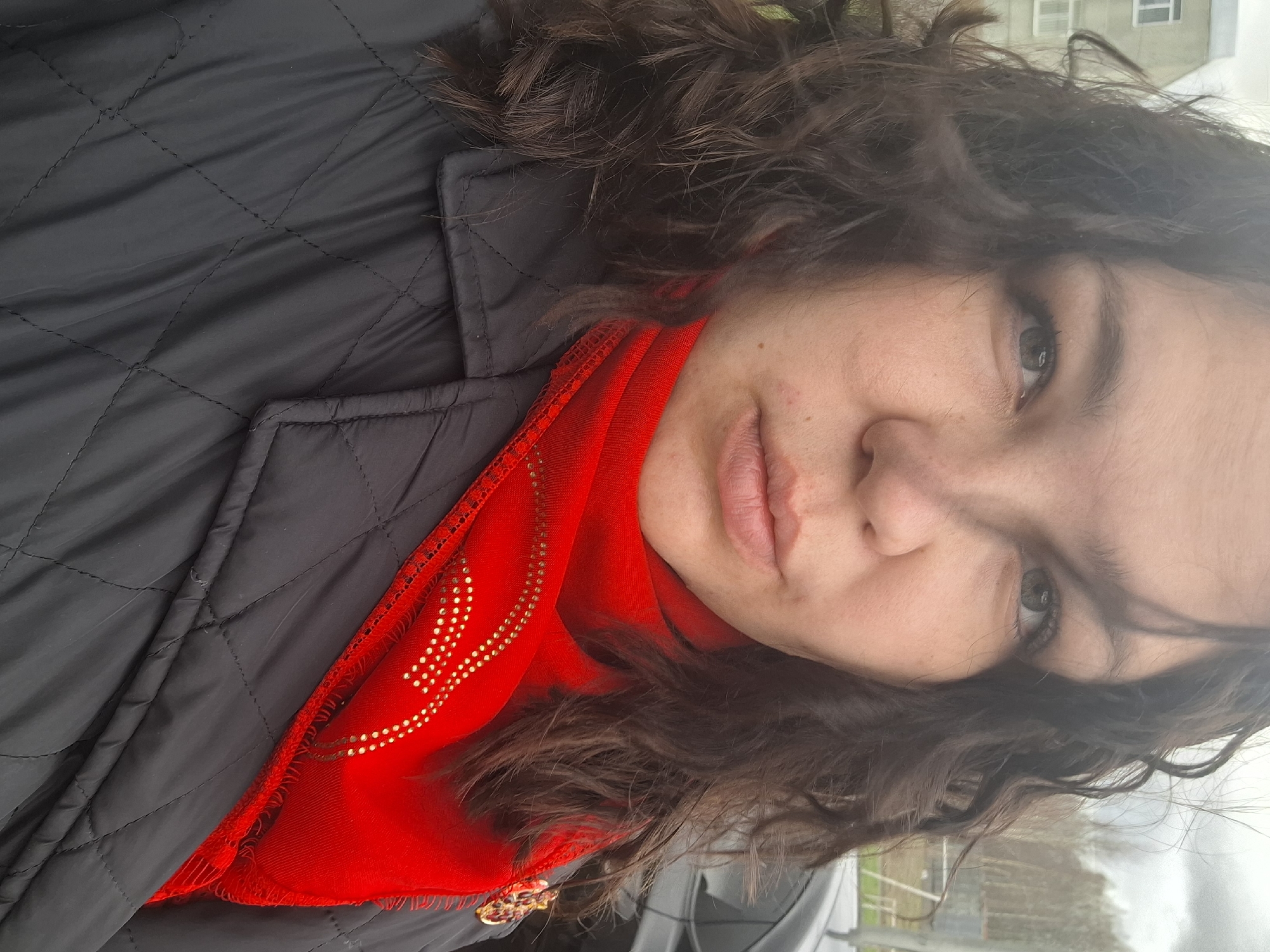 This media file presents a close-up, rotated portrait of a person, likely female, situated outdoors in Марковский, Russia. The individual has dark, curly or wavy hair and looks directly at the viewer with light-colored eyes and a neutral expression. Their skin is fair, with some minor blemishes visible.
They are dressed in a black, quilted jacket with a visible stitched pattern. A vibrant red scarf or shawl is worn beneath the jacket, prominent across the chest and neck. This scarf is adorned with a decorative pattern of numerous small, golden dots, arranged in stylized, non-legible motifs that resemble flowing lines. The scarf also exhibits fringed edges.
The background is softly blurred, indicating an outdoor setting, likely on an overcast day due to the diffused lighting. Faint details in the background include parts of a light-colored building with windows, and indistinct shapes that could be trees, fencing, or a vehicle. The image captures a direct, personal shot, possibly a selfie. There is no legible text visible, only the decorative golden dot pattern on the red scarf.