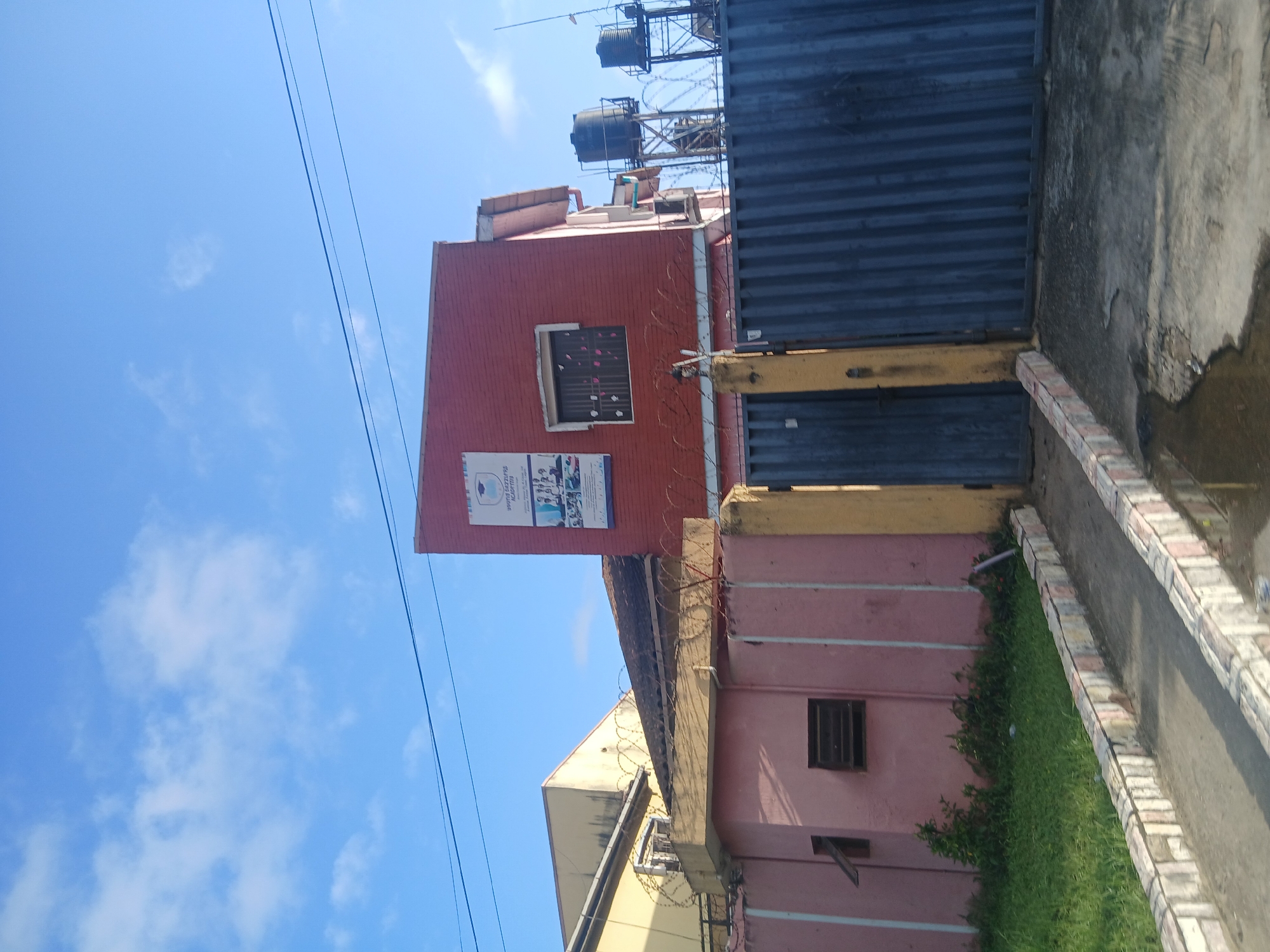 A tall, narrow, pink building with an attached yellow building sits behind a metal gate in Isunba, Nigeria. A red building with a sign showing a graduation cap and students stands to the right of the pink building. Two water tanks on metal stands are visible behind the red building. The sky is blue with scattered white clouds.