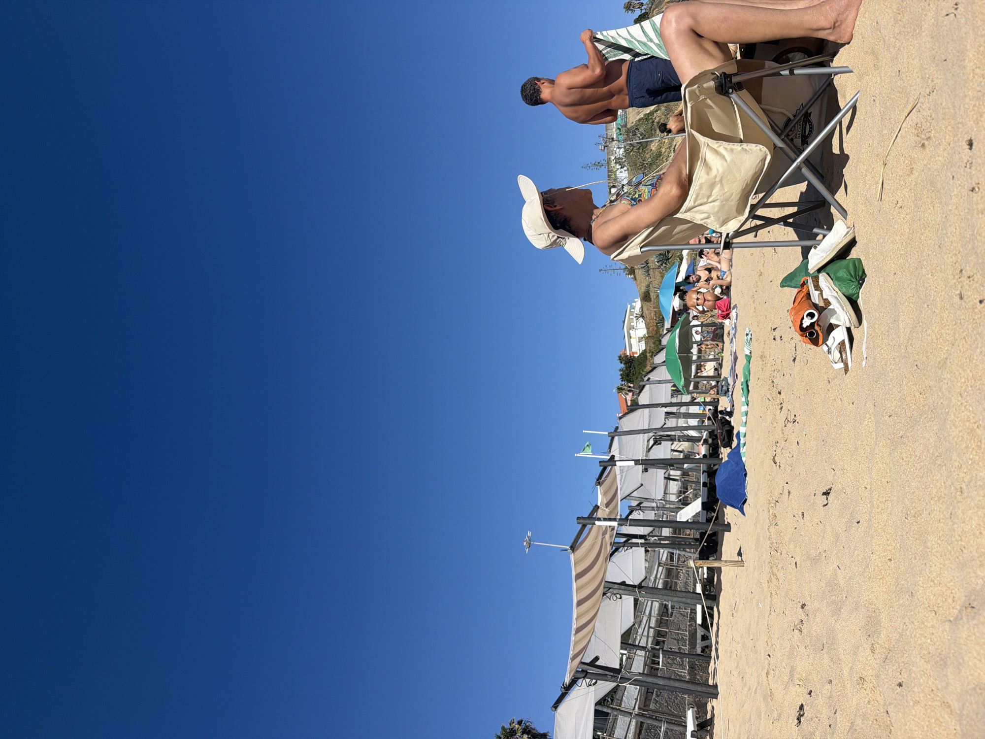 The media file depicts a sunny day at a beach, likely in Cascais, Portugal, based on the location context. The scene is dominated by a clear, deep blue sky, with a vast expanse of sand occupying the right side of the frame. Several people are visible enjoying the beach.
In the foreground, a person wearing a white hat and a patterned shirt is seated in a beige folding chair, looking to the left. Their legs and feet are visible, and they are wearing a necklace. Next to the chair, on the sand, are a pair of white espadrilles, a green folded item, and a pair of sunglasses with orange lenses. Behind this person, another individual is standing, holding a striped towel or blanket. This person is wearing dark blue swim trunks.
Further back, a collection of beach umbrellas and shaded areas are set up, with more people visible relaxing or sunbathing. The structures supporting the umbrellas appear to be made of wood and metal. The sand is light brown and textured, with some scattered debris.
The overall impression is one of a relaxed and pleasant day at the beach. The bright sunlight and clear sky suggest fair weather, likely midday. The people appear to be enjoying their leisure time. There is no visible text in the media file.