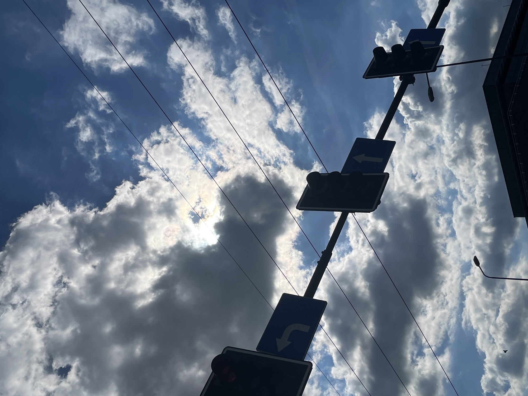 The image shows a low-angle view of a street in an unknown city. A traffic light pole with multiple lights and direction signs dominates the frame, silhouetted against a bright, cloudy sky. Power lines run across the sky. The silhouette of a building is visible on the right side of the frame.