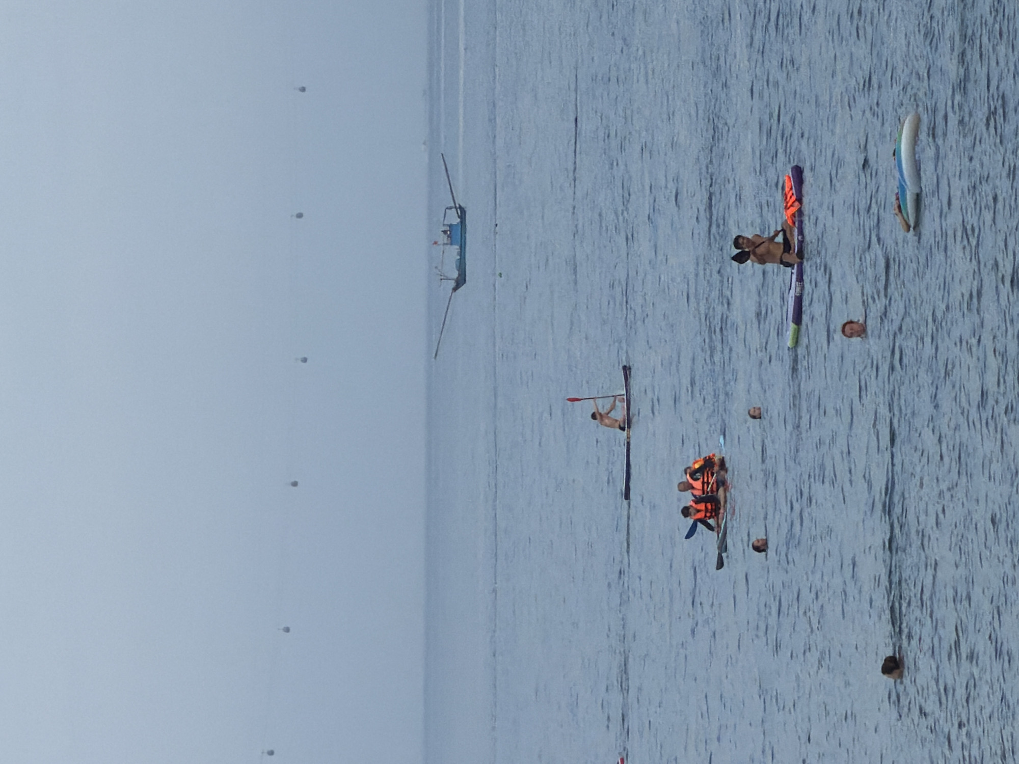 The image depicts a coastal scene in Nha Trang, Vietnam, on a hazy day. The sea is calm and a light blue, with a distant horizon. In the foreground, several people are engaged in water activities. Three individuals are swimming, with only their heads visible above the water. Two more people are on a paddleboard; one is a male wearing an orange life vest and holding a paddle, while the other is a female also wearing an orange life vest. Further back, another paddleboard carries two people, both wearing orange life vests. To the left, a person is on a small red object in the water. In the mid-ground, a single person is on a paddleboard, facing away from the camera. Further in the distance, a blue fishing boat with a large outrigger is sailing. In the far distance, several small, dark objects are visible suspended in the hazy sky, likely part of a cable car system. The overall atmosphere is serene, with a muted color palette due to the overcast conditions.