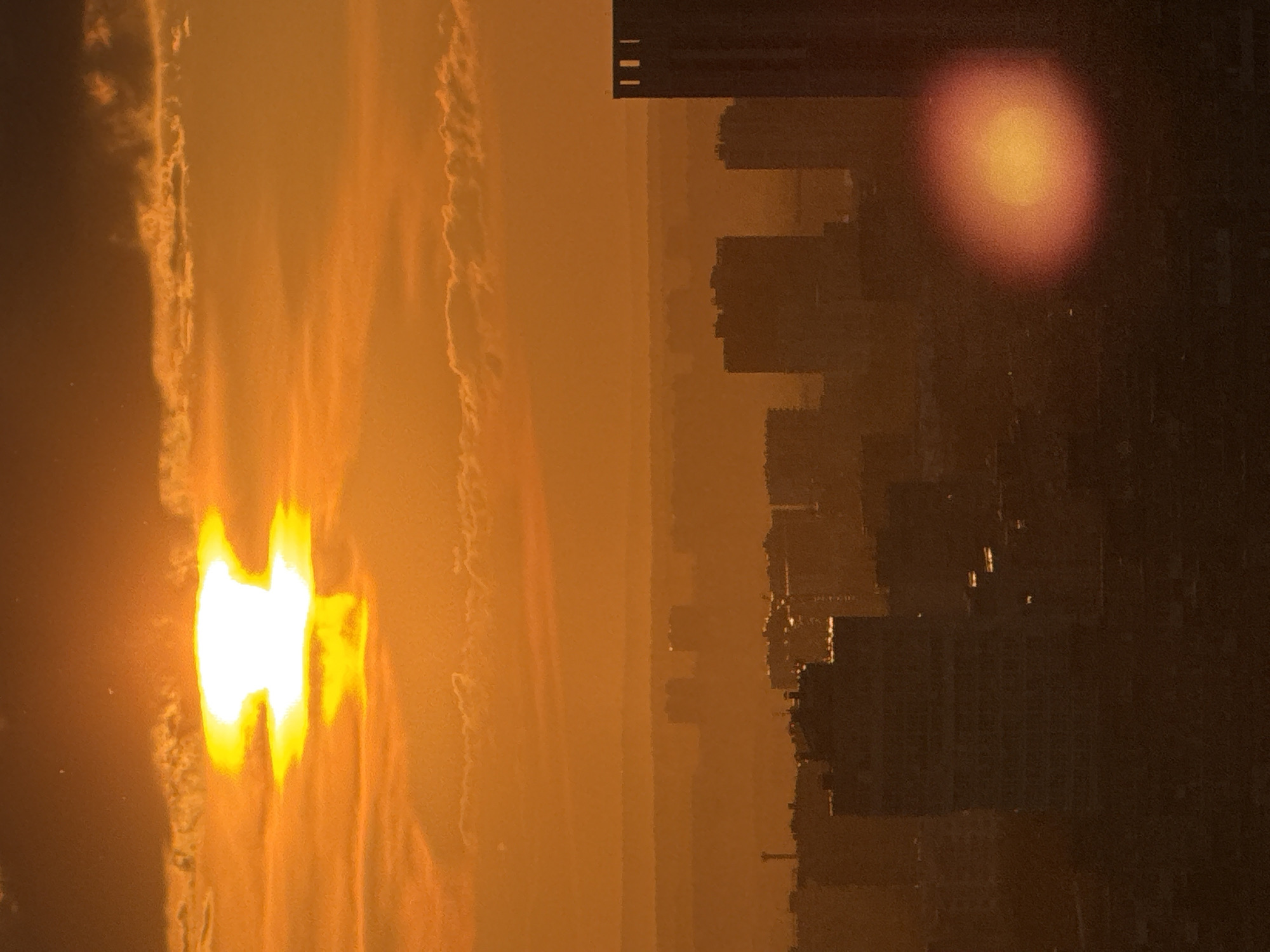 This is a vertical, medium-shot photograph of a city skyline at sunset, rendered in warm, orange hues. The sun, a bright, blown-out white disk with an orange halo, dominates the upper left quadrant of the frame, partially obscured by thin, wispy clouds. The sky transitions from a dark, burnt orange at the top to a lighter, hazy orange towards the horizon.
Below the sky, multiple layers of the cityscape are visible, diminishing in clarity with distance due to atmospheric haze. Silhouetted buildings of varying heights and shapes form the urban landscape. In the foreground, a large, dark building occupies the lower right portion of the image, its top featuring a distinct rectangular structure with three vertical indentations. To the left and center, several other buildings are visible, appearing as dark masses against the bright sky. A prominent, somewhat squat building with a flat roof is centered in the lower half of the image. In the mid-ground, more indistinct buildings are seen, and in the far distance, faint outlines of even more distant structures are discernible. A lens flare, appearing as an oval-shaped, bright orange-yellow light source, is positioned in the lower right quadrant, overlaying the cityscape.
The image is undated and lacks specific context, however, based on the architectural styles and general appearance of the city, it is highly suggestive of an urban environment in Russia, and specifically could be interpreted as an impression of Perm, Russia due to the common presence of such high-rise apartment blocks and the atmospheric conditions often observed during sunsets over industrial or densely populated areas in Russian cities. The overall impression is one of urban sprawl bathed in the dramatic light of a setting sun, creating a somewhat melancholic or awe-inspiring atmosphere. There are no people visible in the photograph.