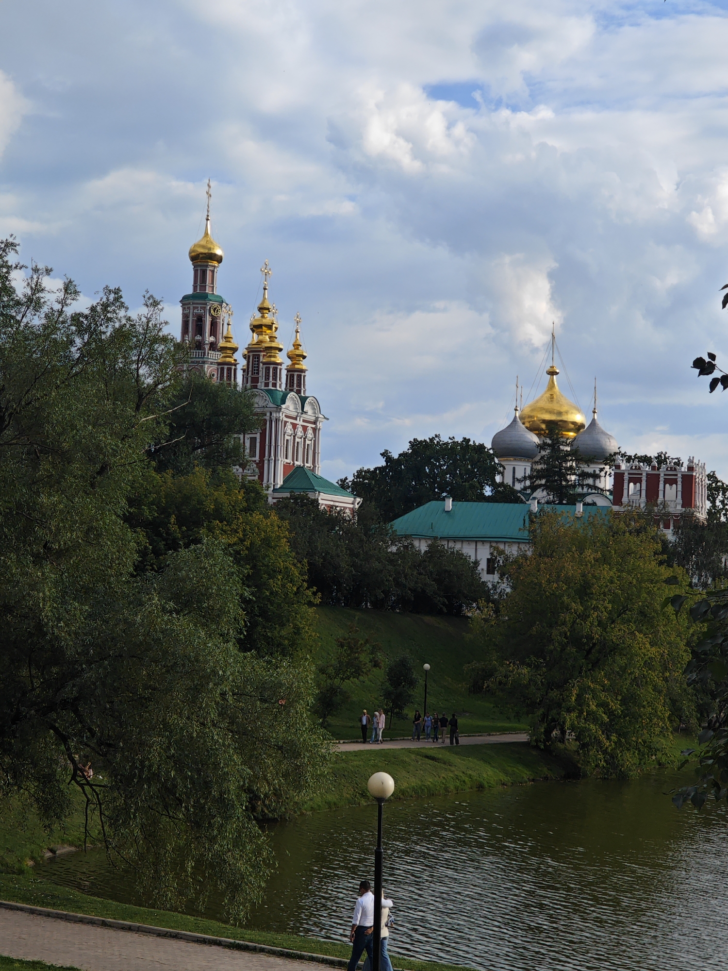 This image shows the Novodevichy Convent in Moscow, Russia, on a cloudy day. The convent is a UNESCO World Heritage site and a prominent example of Russian architecture. The foreground features a tranquil body of water, possibly a pond or river, with ripples on its surface. A paved walkway runs along the water's edge, where two people are walking, one in a white shirt and the other in a light-colored jacket. Further along the walkway, on the grassy bank, a group of five people are standing and walking, appearing to be tourists.
Dominating the background is the magnificent Novodevichy Convent, with its distinctive red and white facade and multiple golden domes, several of which are topped with crosses. The convent's architecture includes various towers and buildings, with green roofs visible on some structures. Lush green trees and foliage surround the convent and line the banks of the water, contributing to the serene and picturesque atmosphere of the scene. The sky is a mix of blue patches and dramatic, fluffy white clouds, creating a dynamic contrast with the buildings and nature. The overall impression is a blend of historical grandeur and natural beauty.