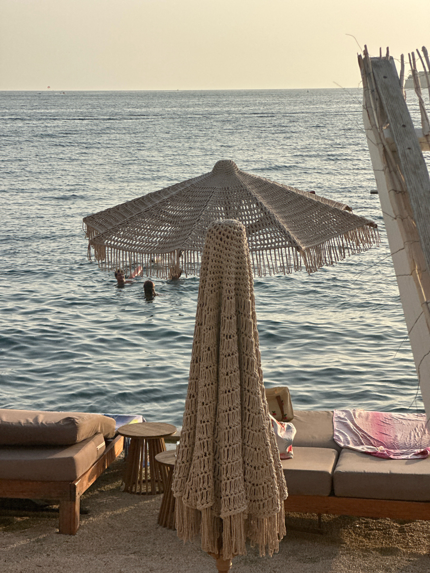 This is a photograph taken in the city of Budva, Montenegro, on a beach. In the foreground, there is a closed thatched umbrella with a macrame cover, and to the left of it, a wooden lounge chair with a brown cushion. Further back, a person is visible partially submerged in the sea. The sea itself is calm with gentle ripples, and the sky is a pale, hazy yellow, suggesting either sunrise or sunset. In the distance, a few boats are visible on the water, and on the right side of the image, there is a white structure with wooden elements, possibly part of a beach bar or cabana. The scene conveys a sense of relaxation and leisure at a coastal resort.