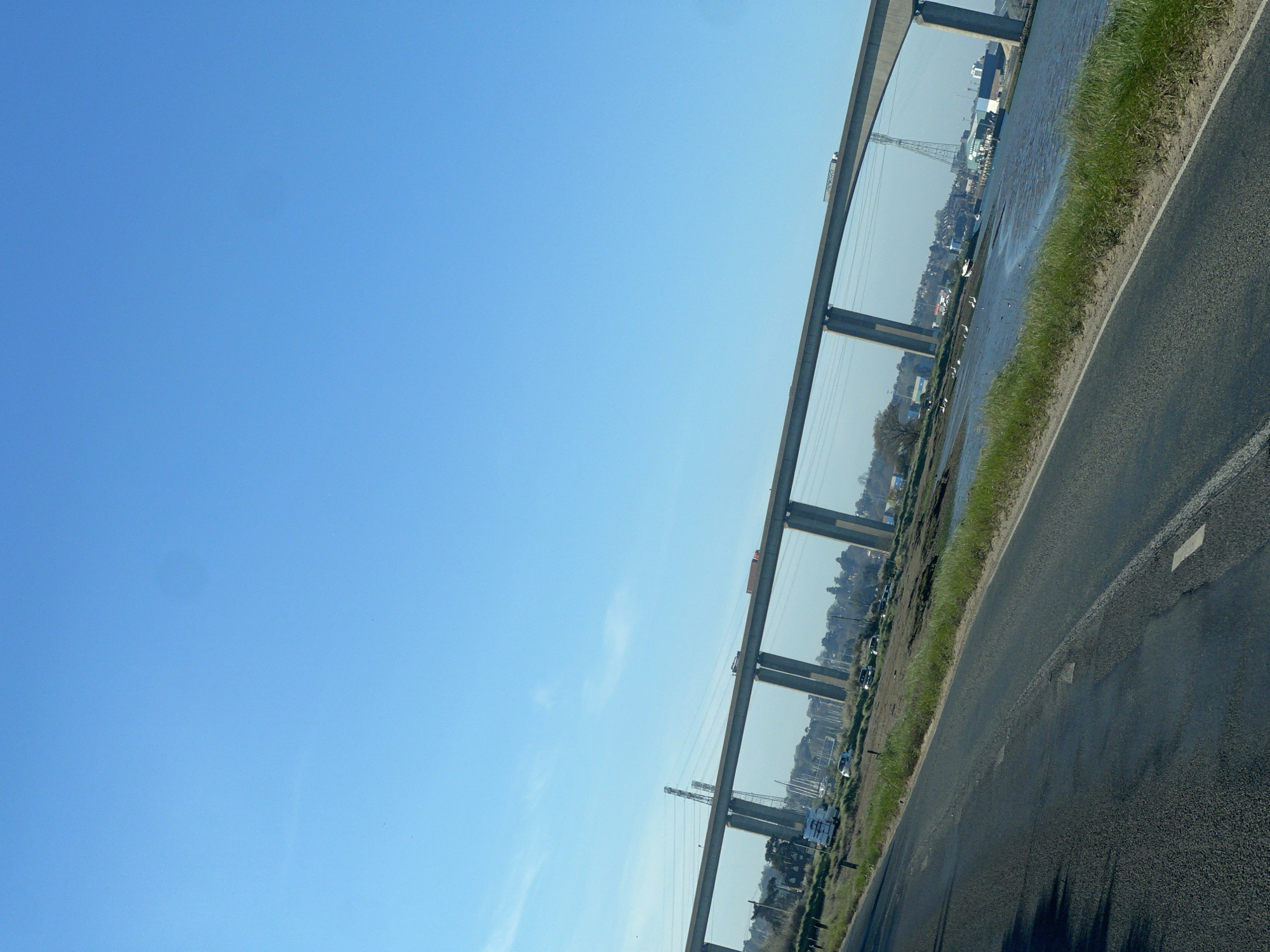 The image depicts a road running alongside a body of water, possibly a river or an estuary, under a bridge structure. The scene suggests a rural or semi-rural setting. Across the water is a settlement with buildings, trees, and what appears to be industrial or port infrastructure, including transmission towers. The sky is clear blue. The location is consistent with a landscape in Babergh, United Kingdom, characterized by waterways and bridges near urban areas. There are no people visible in the image.