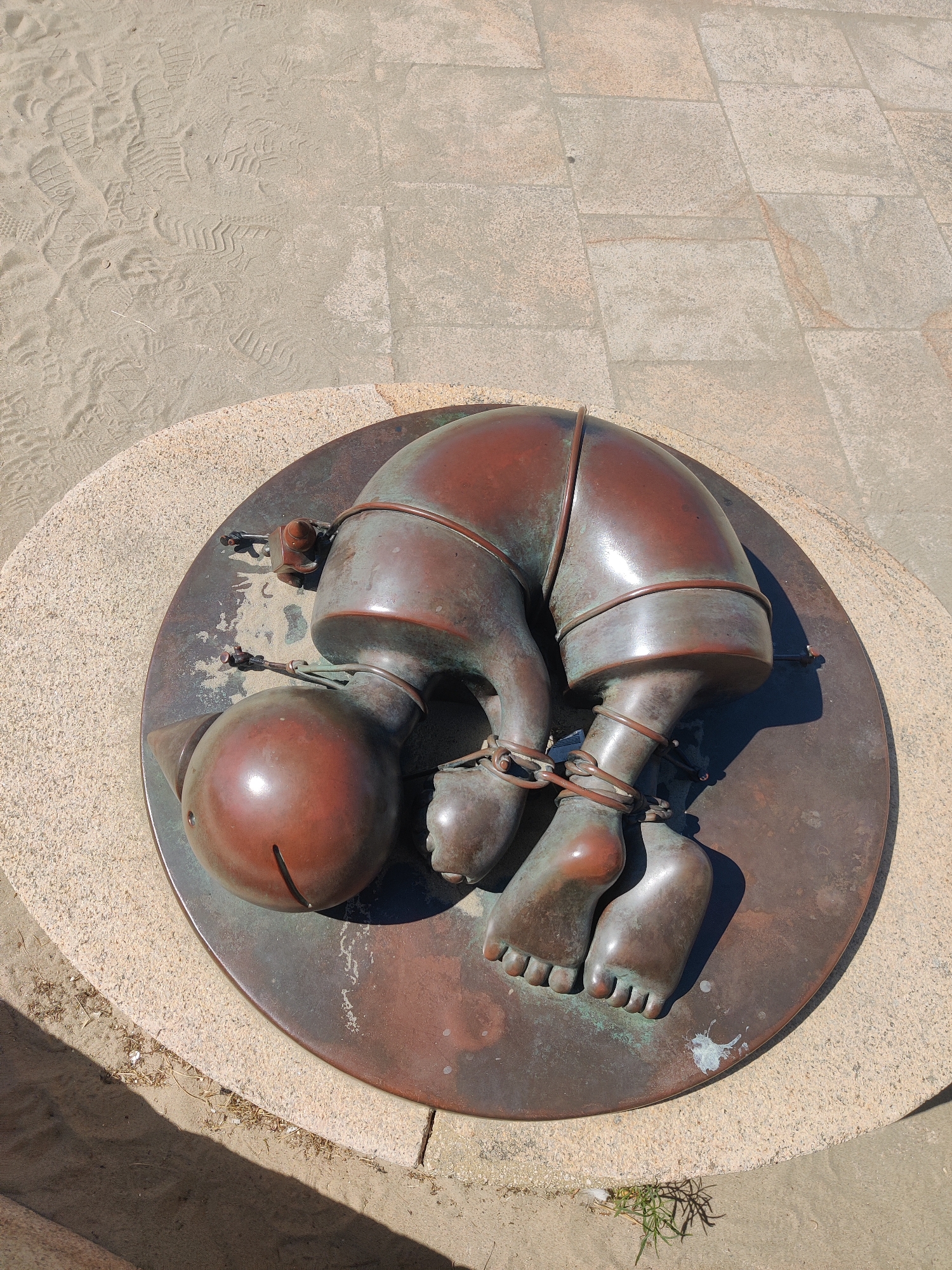 The image shows a bronze sculpture of a person's legs, feet, and head curled into a fetal position, bound by chains. The sculpture is resting on a circular bronze plaque, which in turn is mounted on a textured circular stone base. This base is situated on a surface composed of both sand and paving stones. Shoe prints are visible in the sand to the left of the sculpture. The sculpture is located on the Scheveningen beach promenade in The Hague, Netherlands.