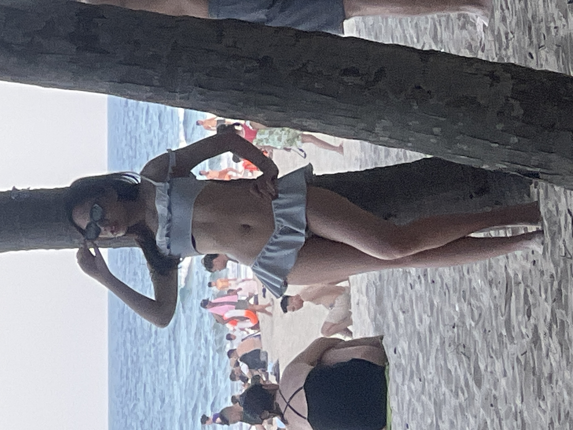 The image depicts a scene at a beach, likely in Da Nang, Vietnam, with the ocean in the background and sand in the foreground. A woman wearing a light blue bikini with ruffled edges is posing against a tree trunk. She has sunglasses on and is touching her hair. Several other people are visible on the beach, some sitting or standing near the water, while others are further back. The water appears to be choppy, indicating waves. The overall lighting suggests it is daytime, possibly late afternoon.