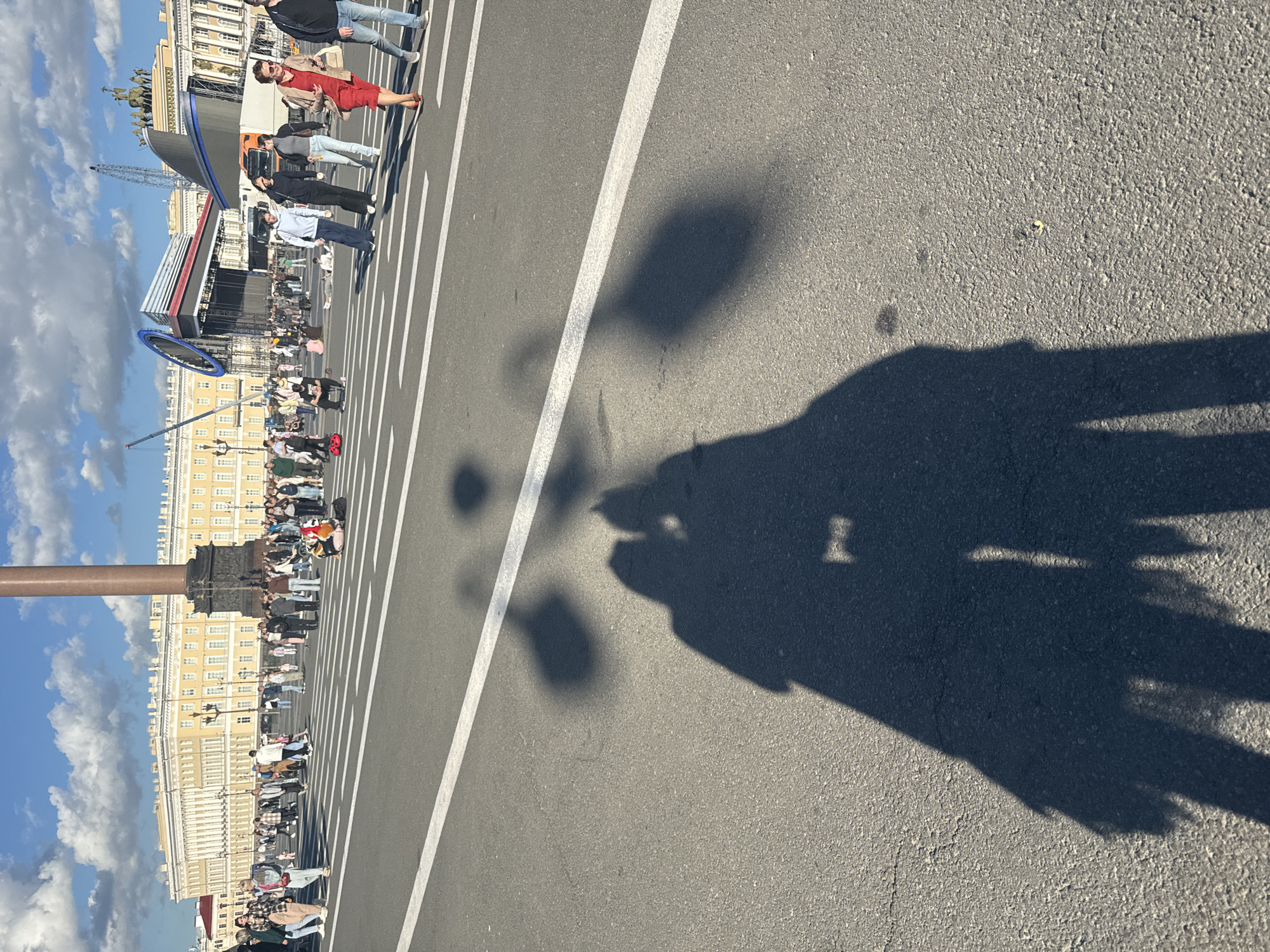 The image depicts a sunny day in Palace Square, Saint Petersburg, Russia, with a prominent Alexander Column in the background. A large stage structure, likely for an event, is set up in the middle ground, with cranes visible behind it. Numerous people are crossing the square and milling about. In the foreground, the long shadows of two individuals are cast across the asphalt, with one shadow appearing to hold a flag or banner. The ground is marked with white pedestrian crossing lines. The sky is blue with scattered white clouds.
