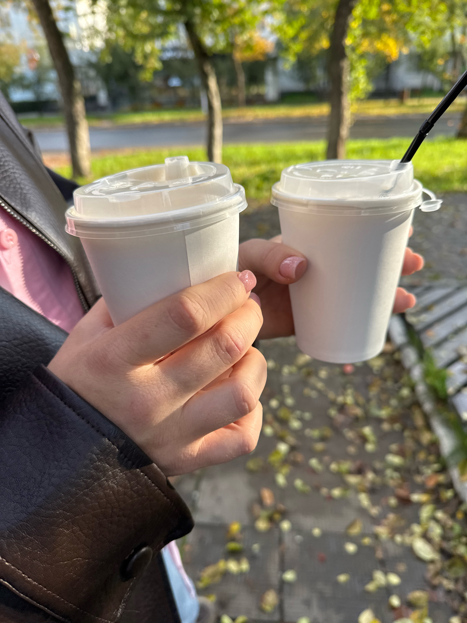 The image captures a close-up, outdoor scene in Chaikovsky, Russia, focusing on two hands holding white paper coffee cups with clear plastic lids. The hands, belonging to a person wearing a dark brown leather jacket over a pink garment, are positioned centrally in the frame. One hand is holding a cup on the left, while the other hand, to the right, is holding a second cup with a black straw protruding from its lid. The background is softly blurred, revealing trees with autumn foliage, a paved area with scattered fallen leaves, and hints of a building and road, suggesting a park or street-side setting. The lighting indicates a daytime, possibly sunny, environment, with shadows cast on the cups and hands.