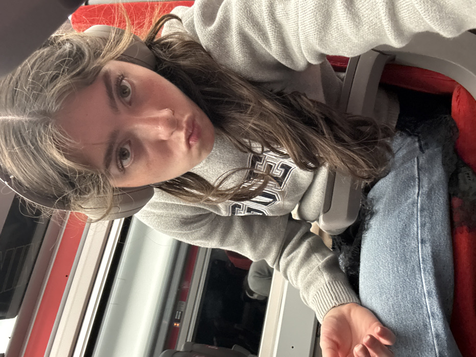 A young woman wearing a gray sweatshirt with a collegiate logo, blue jeans, and headphones poses for a selfie. She has light brown hair and a nose ring. The image is taken at an angle, showing the interior of what appears to be a train car. Red seats and a window can be seen in the background. The woman is making a slight "duck face" expression.