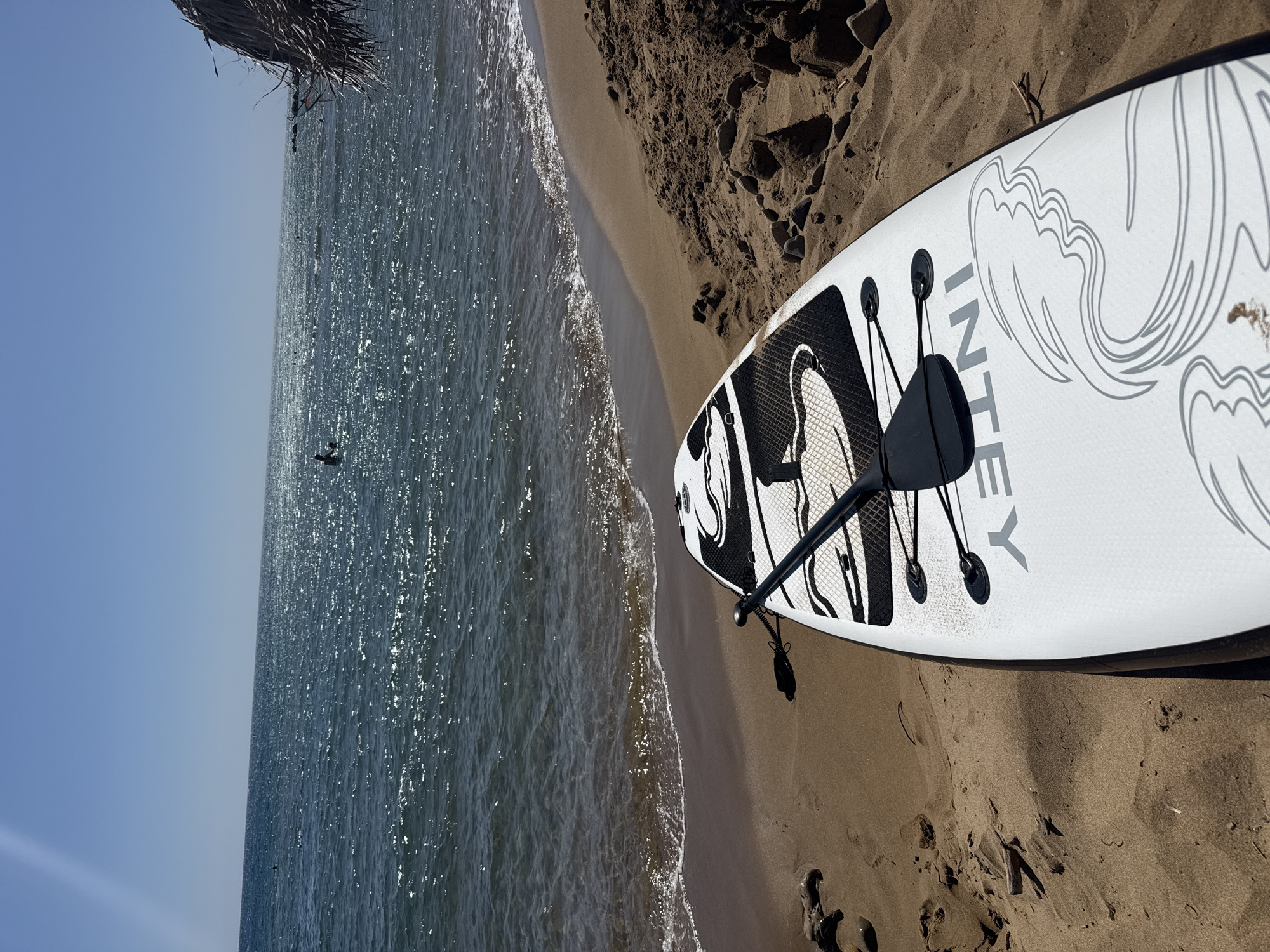 A close-up shot shows a stand-up paddleboard and paddle resting on a sandy beach. The paddleboard is white with black and grey wave-like graphics and the brand name "INTEC" is visible. The paddle is black and secured to the board with bungee cords. The beach is made of golden sand and pebbles, with small sand formations in the foreground. The sea is calm and sparkling under the bright sun, with a few distant figures visible in the water. The sky is clear blue. The scene is located in the Municipal Unit of Lefkimmi, Greece, likely on a beach in a coastal area.