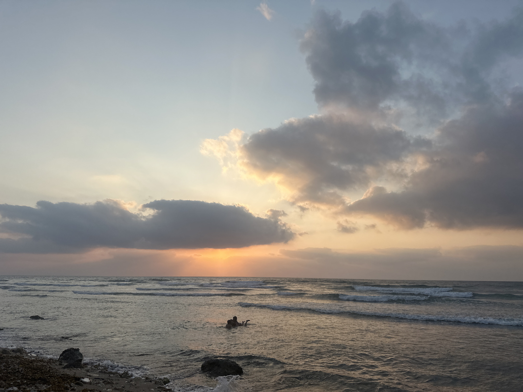 The image shows a wide, sandy beach with a gentle surf. The sky is a soft blue, with a few scattered clouds tinged with the colors of a sunset. The sun, which is partially obscured by clouds, casts a warm glow over the scene. In the foreground, a few rocks and some seaweed are visible on the shore. In the water, a person is seen swimming, with their head and shoulders above the surface. The overall impression is one of peace and tranquility.
This beach is located in Haifa, Israel.