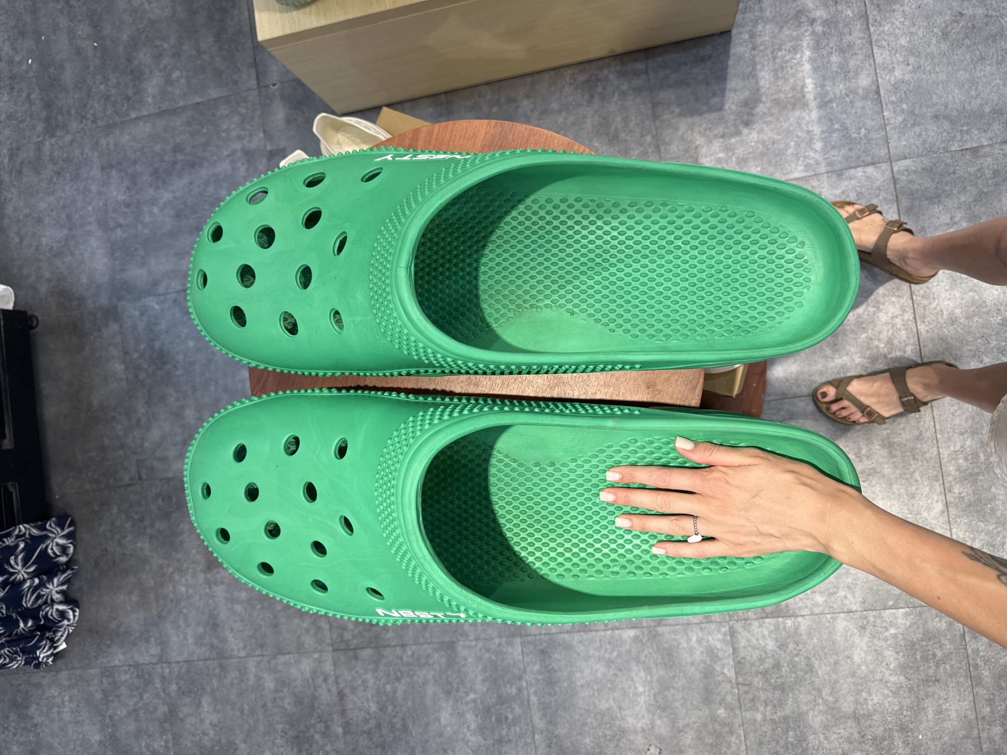 A person's hand with manicured nails and a ring is shown placed inside a very large, bright green clog shoe. The shoe is one of a pair, both of which are visible in the image, also very large and bright green. The clog has the characteristic holes on top and a textured sole. The person's arm is also visible, extending from the hand down the side of the shoe. The scene appears to be indoors, with a grey tiled floor visible around a wooden surface on which the shoes are placed. In the background, part of a dark navy fabric with white palm tree print is visible, along with a light-colored wooden counter. The overall impression is one of extreme scale, highlighting the oversized nature of the footwear. The location is not identifiable within the image.