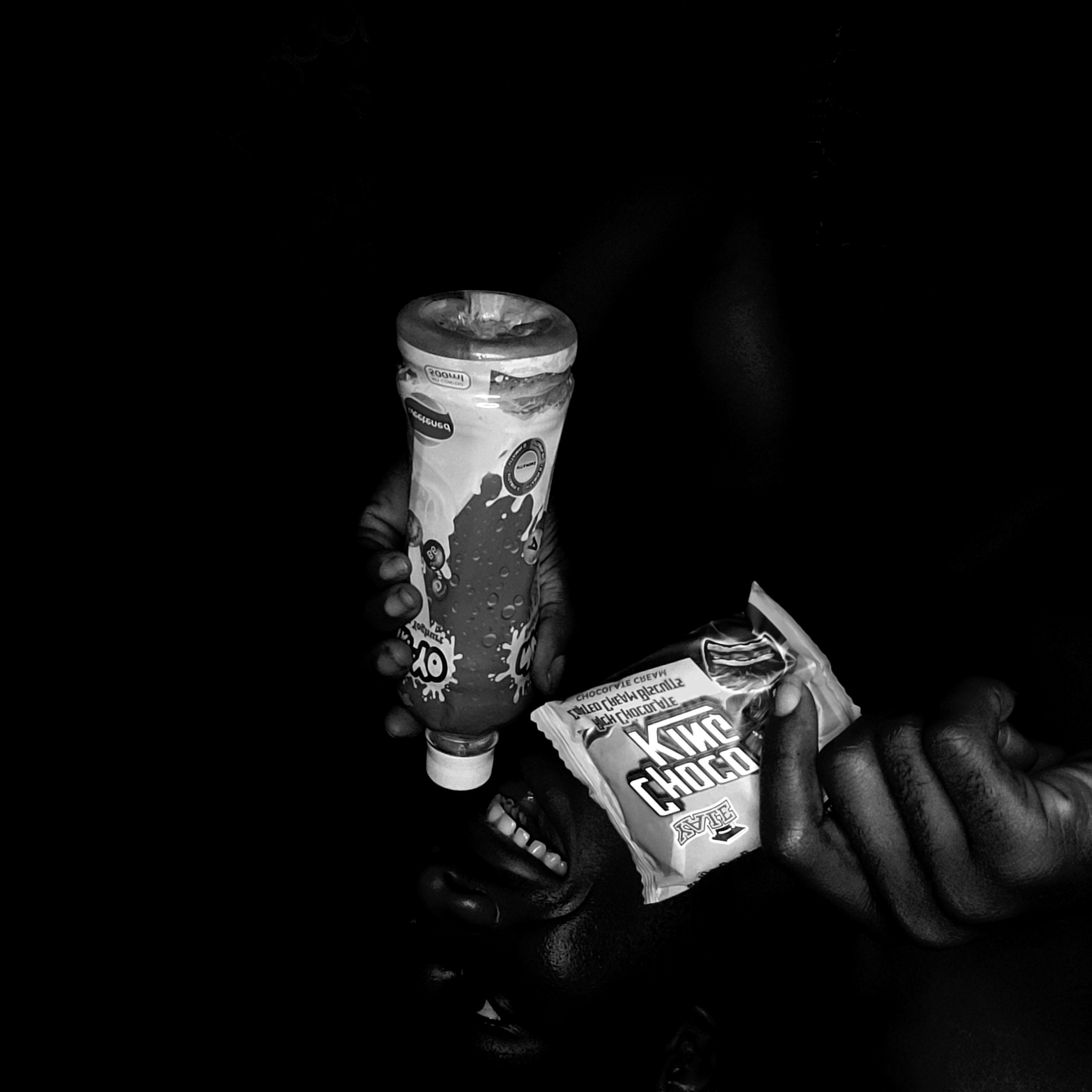 A grayscale photograph reveals a person holding a yogurt drink and a chocolate bar. The hand holding the yogurt drink, labeled "OY Yogurt", is positioned near the top of the image, while the other hand clutches a "King Choco" chocolate bar. The person's mouth is visible, positioned between the two items, and their eyes are visible in the bottom part of the frame. The image is set against a dark background. The photograph was possibly taken in Narayi, Nigeria.