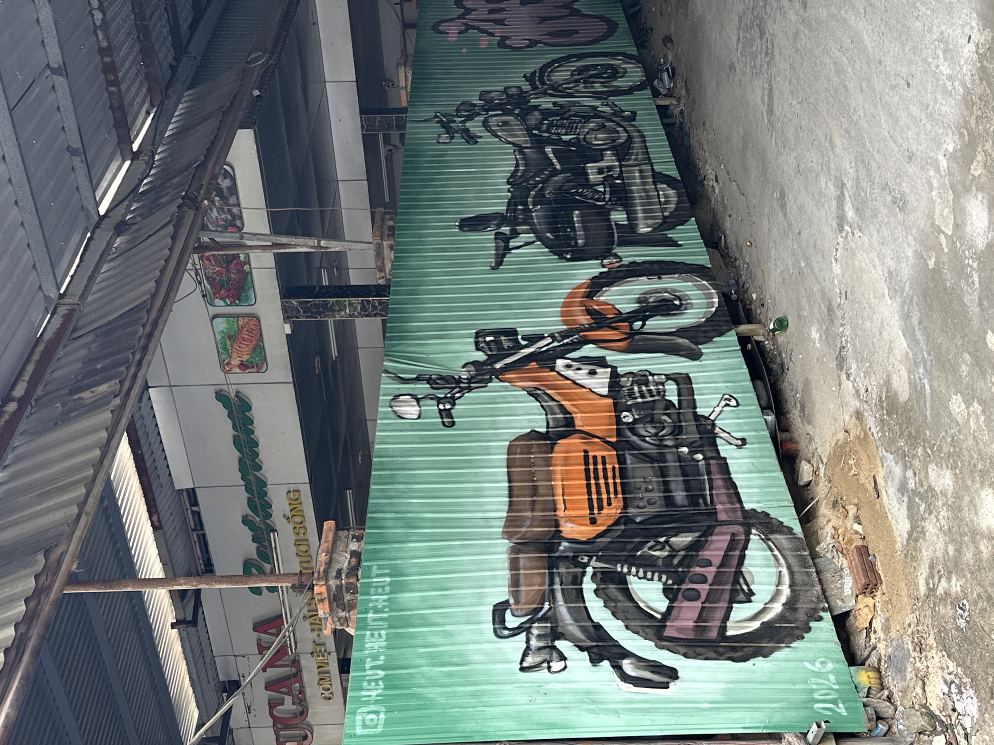 The image depicts a graffiti-covered corrugated metal fence in what appears to be an outdoor market or street setting in Đà Nẵng, Vietnam. The fence, painted a vibrant green, features a mural of two motorcycles. The motorcycle on the left is rendered in orange and black, while the one on the right is in black and white, suggesting a sketch-like style. The mural is detailed, showing the wheels, engines, seats, and handlebars of the motorcycles.
Above the fence, the facade of a building is visible, with signage indicating it was once a restaurant. Part of a sign reads "UCANA" and "Restaurant" in green lettering, with some Vietnamese text below that is partially obscured. There are also framed pictures of food items displayed on the building's exterior.
The environment around the fence is somewhat derelict, with debris and sand on the ground. Bricks and discarded items are scattered near the base of the fence. The overall impression is one of an urban space that is either under construction, renovation, or has fallen into disrepair, with the graffiti serving as a colorful, artistic intervention. The date "2026" is painted in white near the bottom left of the fence.