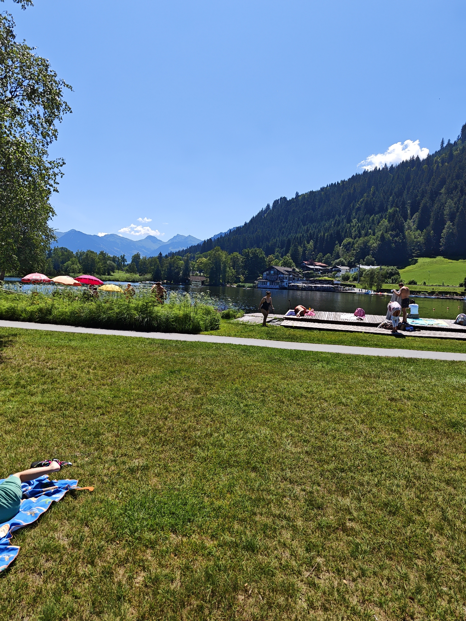 This image depicts a sunny day at a lake in Kitzbühel, Austria. In the foreground, a patch of green grass is visible, with a person lying on a blue towel on the left side. In the middle ground, a lake is present, with a wooden pier extending into the water. Several people are on the pier, some are standing and others are sitting or lying down. There are also several colorful umbrellas near the edge of the lake. In the background, a forested mountain slopes down towards the lake, with some buildings nestled amongst the trees. The sky is clear and blue, with a few white clouds visible in the distance. The overall scene suggests a recreational area during warm weather, with people enjoying outdoor activities.
