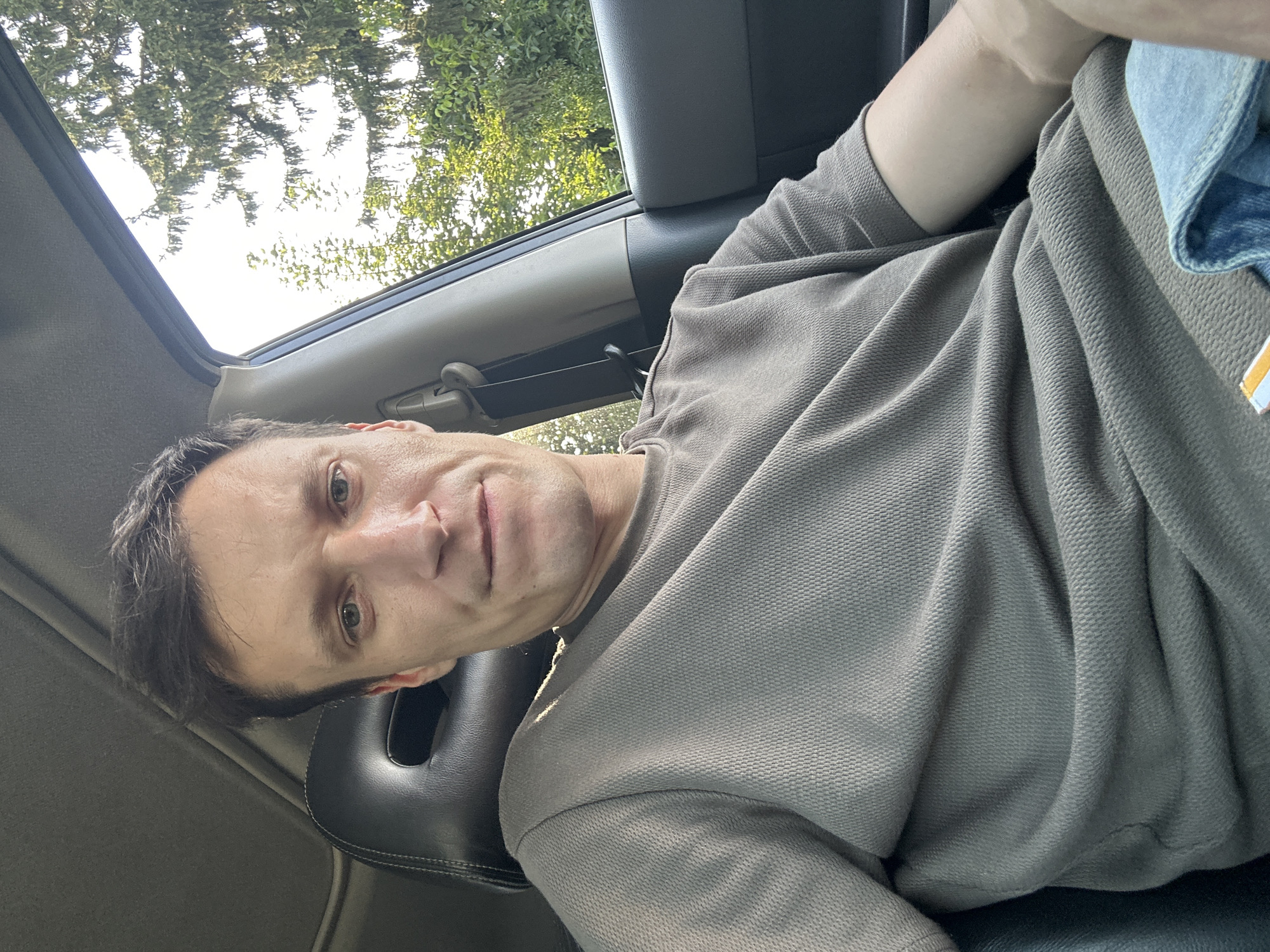 In Shatava, Ukraine, a person is seated in the driver's side of a car, with the camera positioned in front of them. The person is looking directly at the camera. The interior of the car is visible, including the headrest of the seat, part of the dashboard, and a seatbelt. Outside the car, through the open window, trees and a bright sky are visible. The person is wearing a brown t-shirt and blue jeans. The lighting suggests it is daytime.
