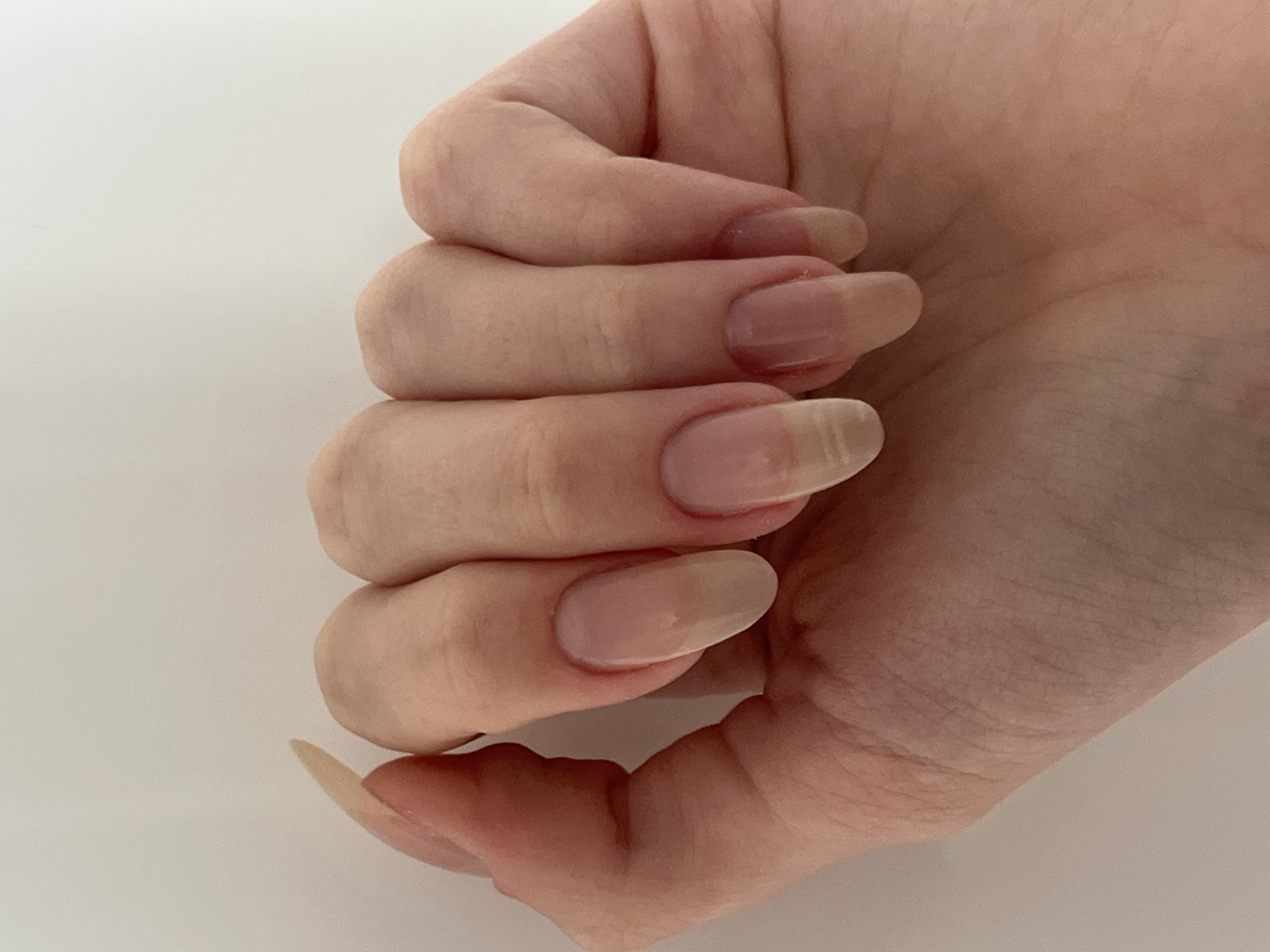 A close-up, top-down view of a person's left hand and fingernails is shown against a plain, off-white background. The hand is positioned with the palm facing downwards and the fingers slightly curled inward. The fingernails are long, oval-shaped, and appear to be clear or have a very sheer, natural-looking polish. The skin of the hand is light-skinned with visible creases and subtle veins. The lighting is soft and diffused, creating gentle shadows. There is no discernible background context indicating a specific location in Chelyabinsk, Russia.