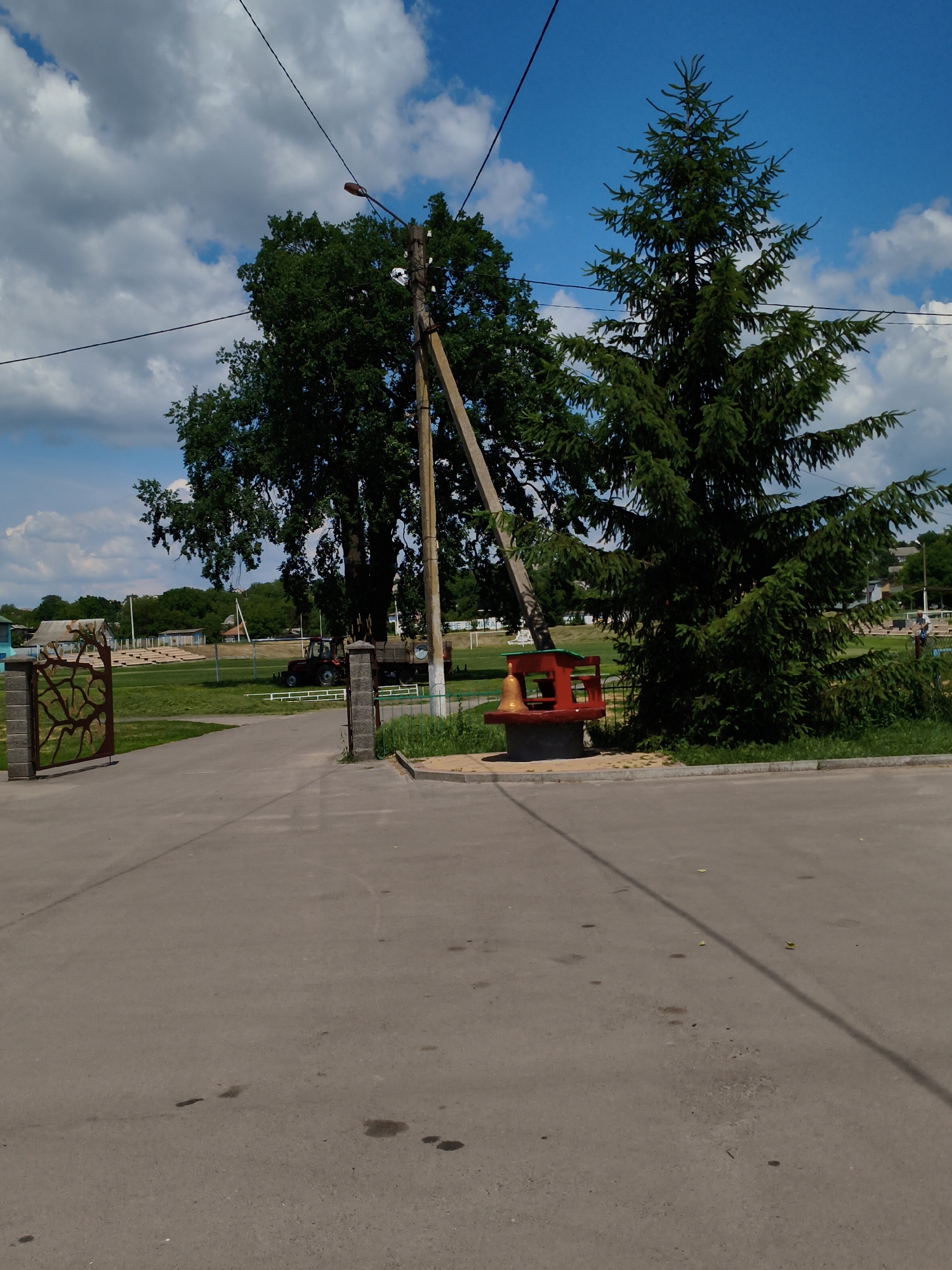 This is a medium shot, outdoor photograph taken on a sunny day in Velyka Bahachka, Ukraine. The scene is a paved road leading into what appears to be a park or recreational area. A large spruce tree dominates the right side of the frame, while a deciduous tree stands to the left behind a utility pole.
In the middle ground, a bright red, bench-like structure with a large golden bell sits next to the base of the spruce tree. Behind the bench, a tractor with a trailer is parked on a grassy area. Further in the background, there are bleachers or stadium seating, suggesting a sports or event venue. A decorative stone gate with an abstract design is visible on the left side of the road, leading into the grassy area.
The sky is blue with scattered white clouds. The overall mood is peaceful and rural. There is no visible text in the image. The time of day appears to be midday, given the bright sunlight and shadows.