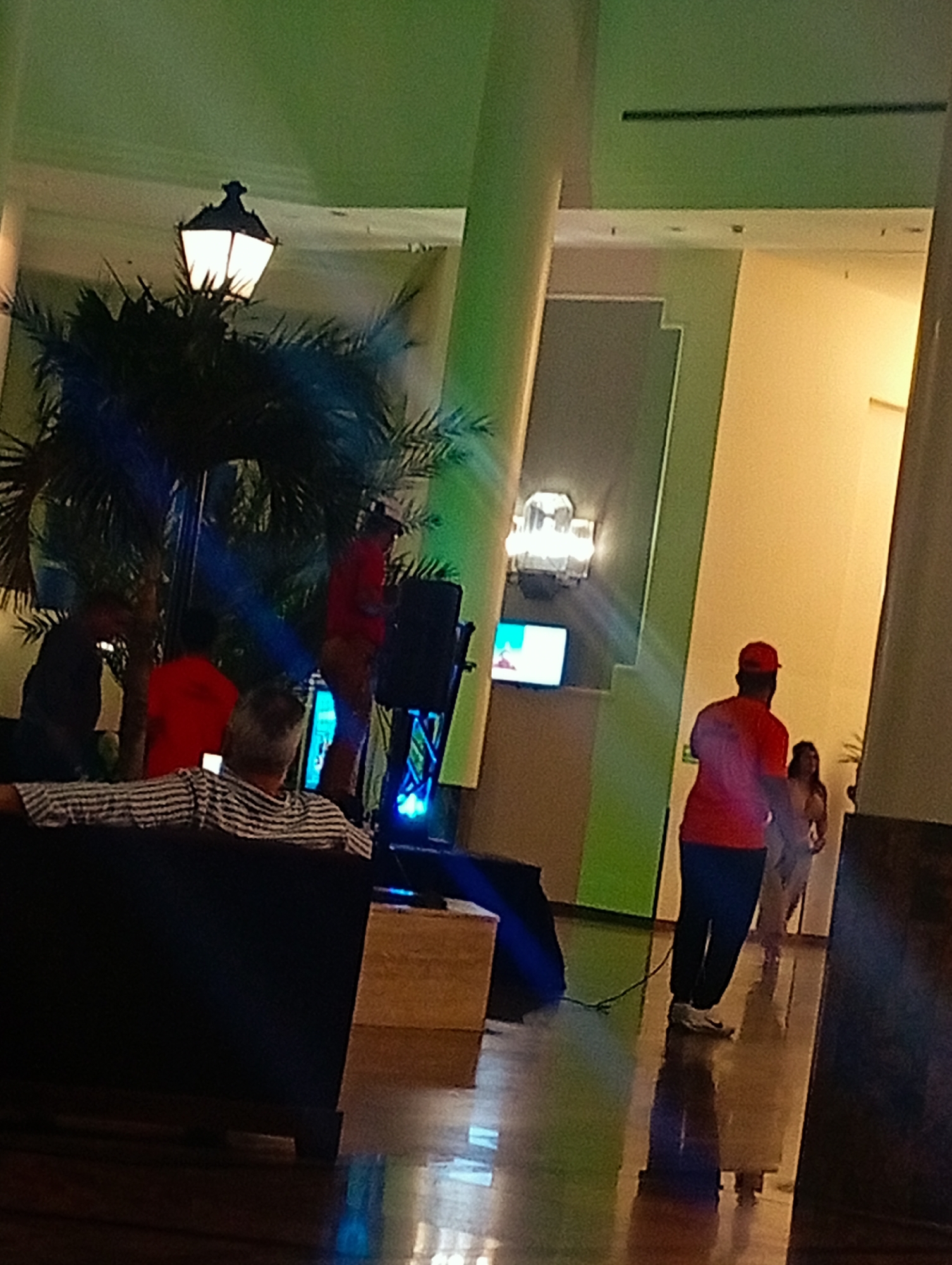The image captures an indoor scene in Pedro González, Venezuela. A group of people is gathered, seemingly for an event or performance. A person in a red shirt and cap is walking away from a small stage area. The stage has a loudspeaker, some type of digital screen and lighting. Two other people are standing on the stage. One is wearing red clothing and the other is wearing dark-colored clothing. Several other people are sitting on a black sofa, watching the stage. A light post with an illuminated lantern can be seen behind the stage. The environment includes shiny hardwood floors and large, decorated architectural columns. The lighting is generally dim, with a green hue on some of the surfaces.