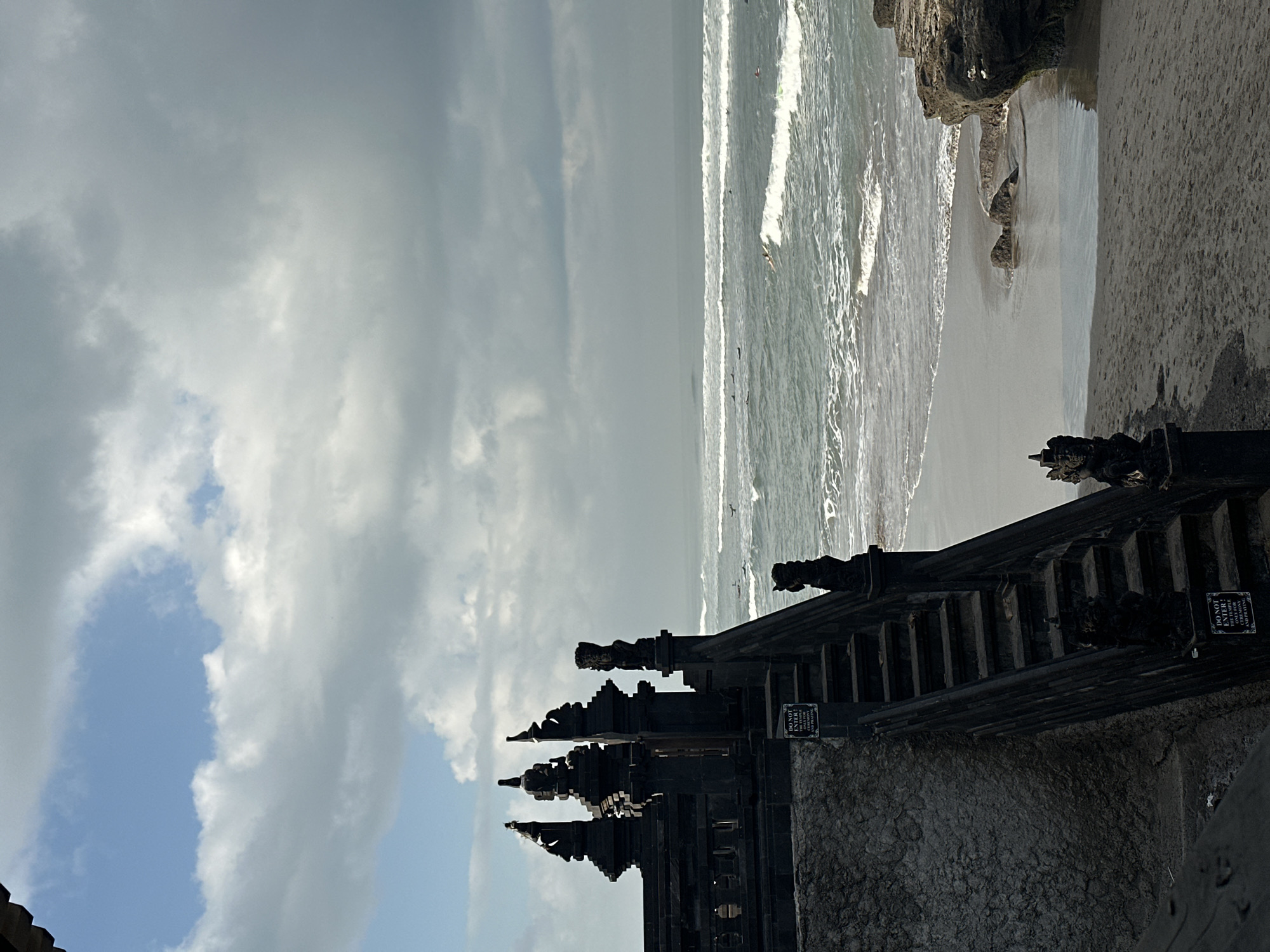 A wide-angle, eye-level shot captures a dark, intricate temple structure with steps leading down to a sandy beach and the ocean. The temple, likely made of dark stone, features elaborate carvings and tiered roofs, silhouetted against a dramatic sky. Several surfers are visible in the water, riding waves that break on the shore. The beach is mostly empty, with some rocks visible near the water's edge. The sky is a mix of blue patches and heavy, gray clouds, suggesting a dynamic weather pattern. The overall mood is serene yet powerful, with the natural elements of the sea and sky contrasting with the man-made temple. This scene is likely located at Tanah Lot Temple in North Kuta, Bali, Indonesia, a well-known sea temple.