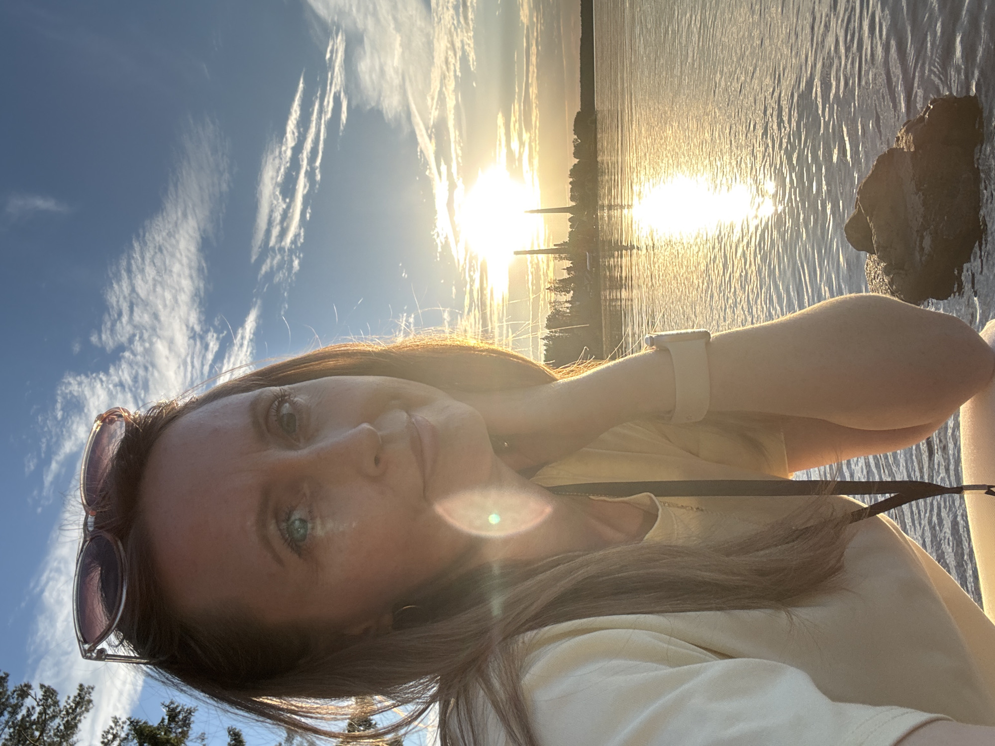 A close-up, selfie-style image captures a woman with long, brown hair and light blue eyes, resting her chin on her hand. She is wearing a light yellow shirt and sunglasses are perched on top of her head. The background features a body of water reflecting the setting sun, with a distant shoreline and a tall, industrial structure. The sky is a vibrant blue with scattered clouds. This scene is set in the city of Chaikovsky, Russia, along the Kama River.