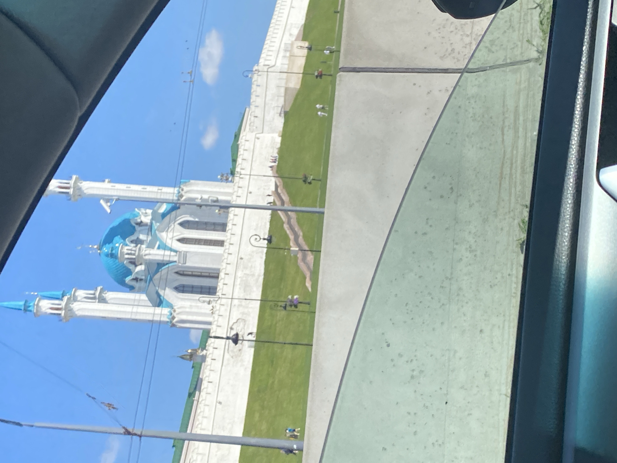 This is a photograph taken from inside a vehicle, looking out at the Kul Sharif Mosque in Kazan, Russia. The mosque is a prominent white building with a bright blue dome and minarets, set against a clear blue sky. The mosque is situated on a grassy hill, with a white wall surrounding its base. Several people are visible on the hill, walking and standing. Streetlights with decorative tops are spaced along the path leading up the hill. The foreground of the image is dominated by the car's interior, including the window and a portion of the dashboard. The overall impression is a sunny day in a historic city.