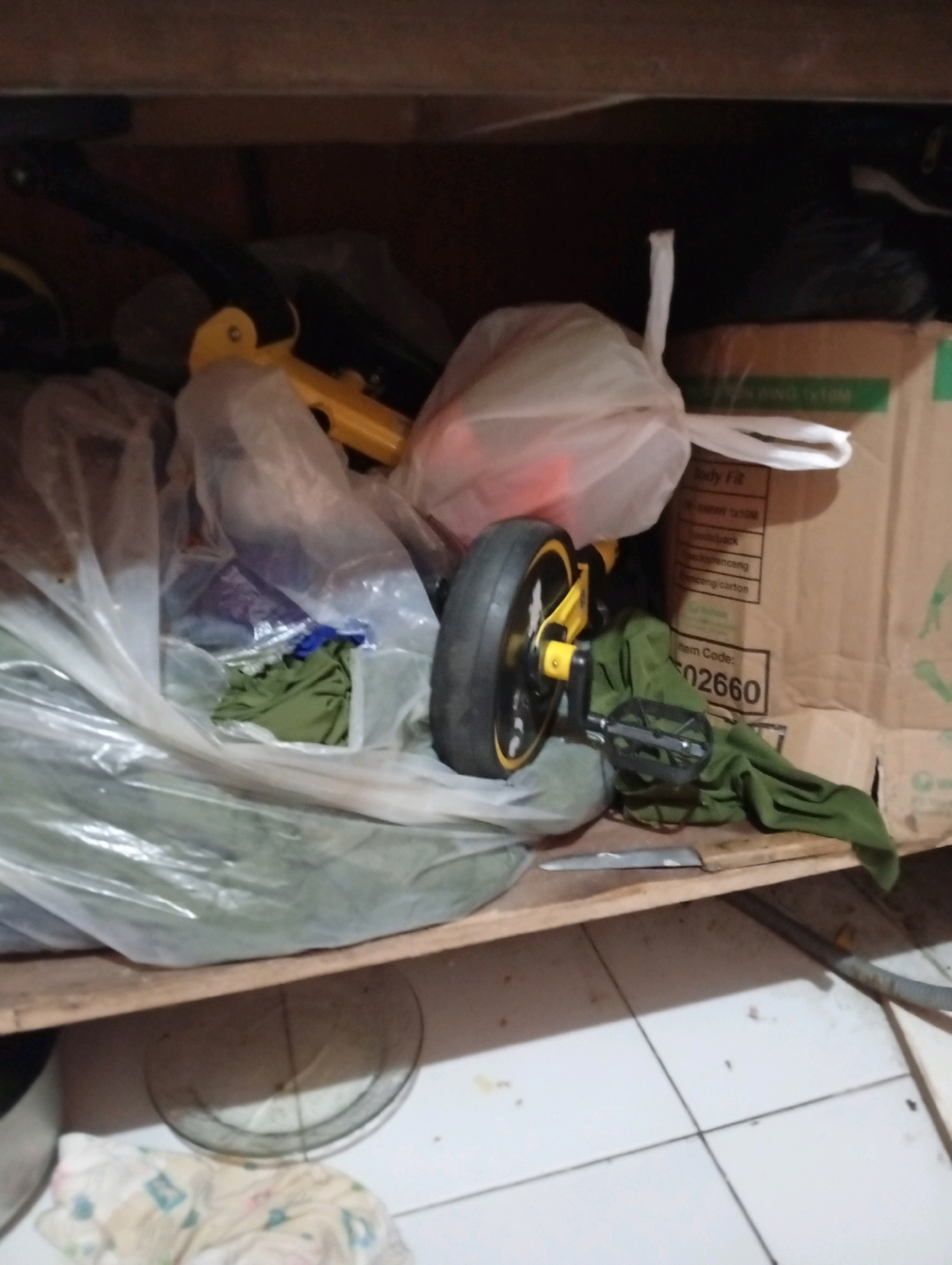 A cluttered shelf in an indoor storage area shows various items. In the foreground, a bicycle wheel with a yellow rim and black tire is prominent, attached to a yellow frame and black pedals. A green cloth is draped over parts of the bicycle and a large clear plastic bag containing more items. Behind the bicycle, a brown cardboard box with green and white text, including "Item Code: 502660," is visible. Other assorted objects, including more plastic-wrapped items and a black handle, are also present on the shelf. The shelf itself appears to be made of wood, and the background shows a dark wooden surface above. The floor below is tiled. There are no people visible in the image, and no specific location in Tasikmalaya, Indonesia, can be determined from the scene.