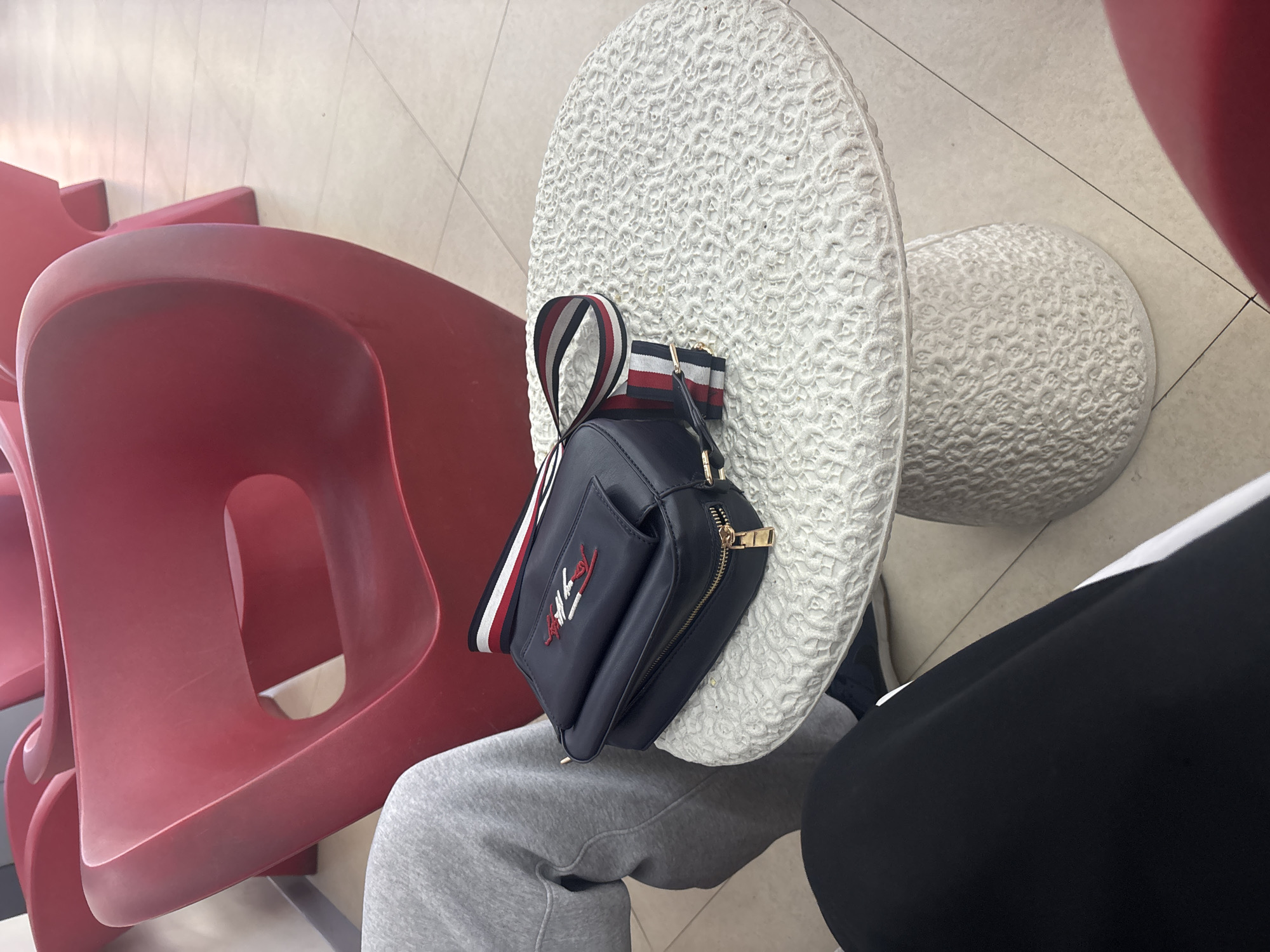 A person is seated in an indoor waiting area, visible from the waist down, wearing grey sweatpants and black and white athletic wear. Their legs are crossed. Beside them, a dark blue (likely navy) cross-body bag with a prominent red, white, and blue striped strap rests on a decorative, white, round-topped stool or side table. The stool features an intricate, textured pattern resembling lace or floral motifs across its surface and base. The bag has a gold zipper and a red and white logo on its front pocket, consistent with a "Tommy" or "Tommy Hilfiger" brand.
In the background and to the left, several red, molded plastic chairs are visible, arranged in a row, suggesting a public waiting area such as an airport, bus station, or a large lobby. The chairs have a modern, sculptural design. The floor is covered with light-colored tiles, and the lighting is bright and even, indicating daytime indoors. The overall setting is a clean, modern public space.