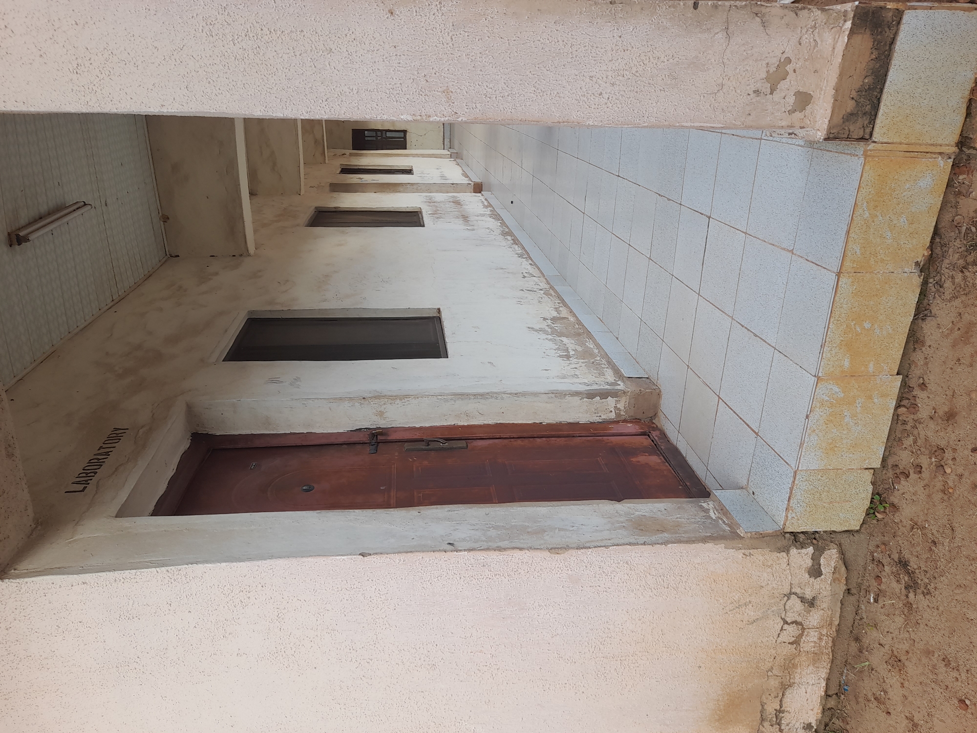 The image shows a hallway of a building. A wooden door with a metal handle is on the left side of the image. Above the door, the word "LABORATORY" is written on the wall. The hallway has a tiled floor and a white ceiling with fluorescent lights. Several windows are visible along the right wall of the hallway. The building appears to be in a state of disrepair, with peeling paint and cracked walls. The ground outside the building is made of dirt and small rocks. The location of this building is in the city of Ibi, Nigeria.
