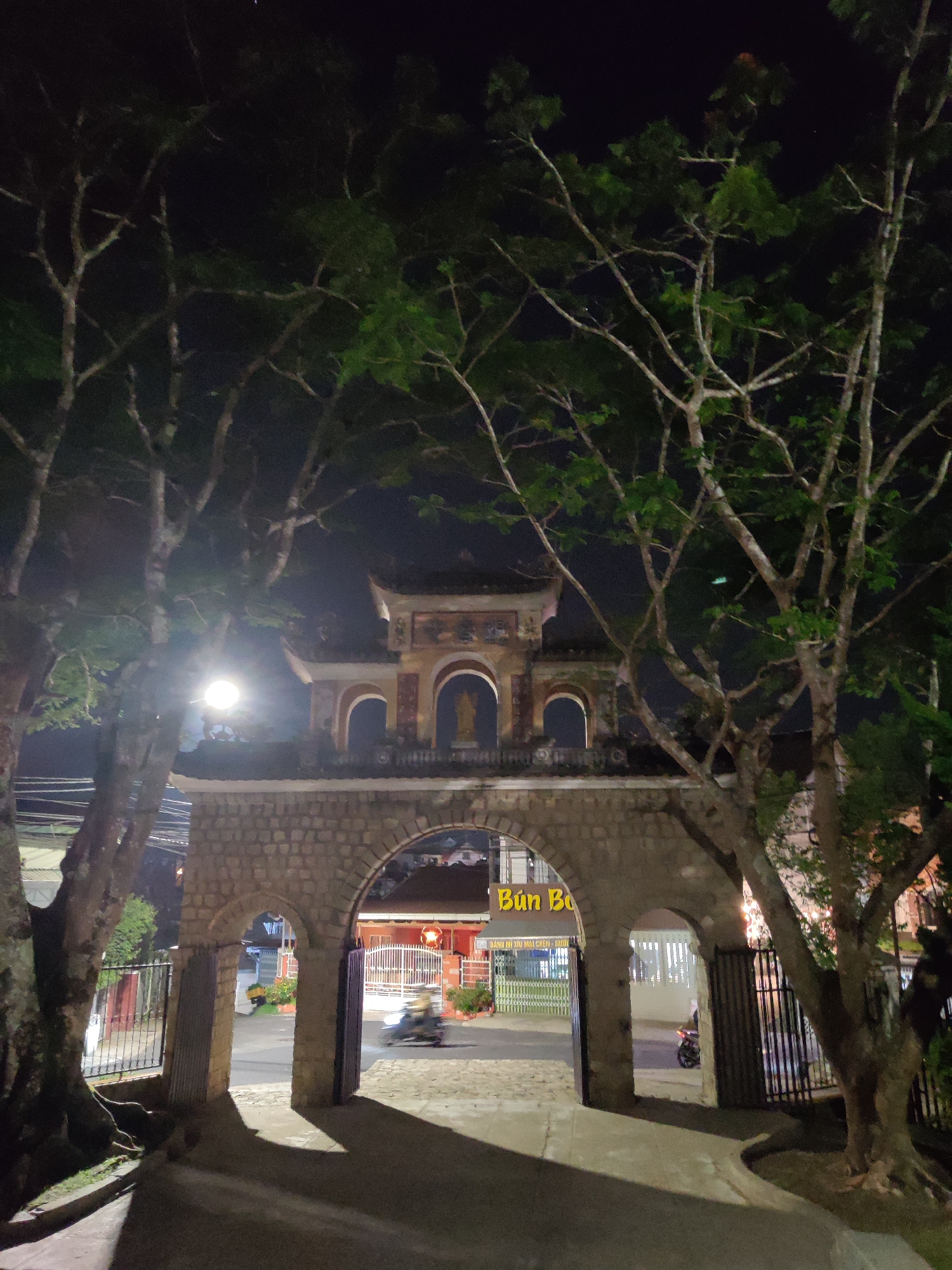 Night view through a stone archway entrance to Tran Hung Dao Street in Da Lat, Vietnam. The archway is flanked by trees. Beyond the archway, a street leads to a building with a sign reading "Bún Bò." A motorcycle is visible on the street. A dark, overcast sky is above.