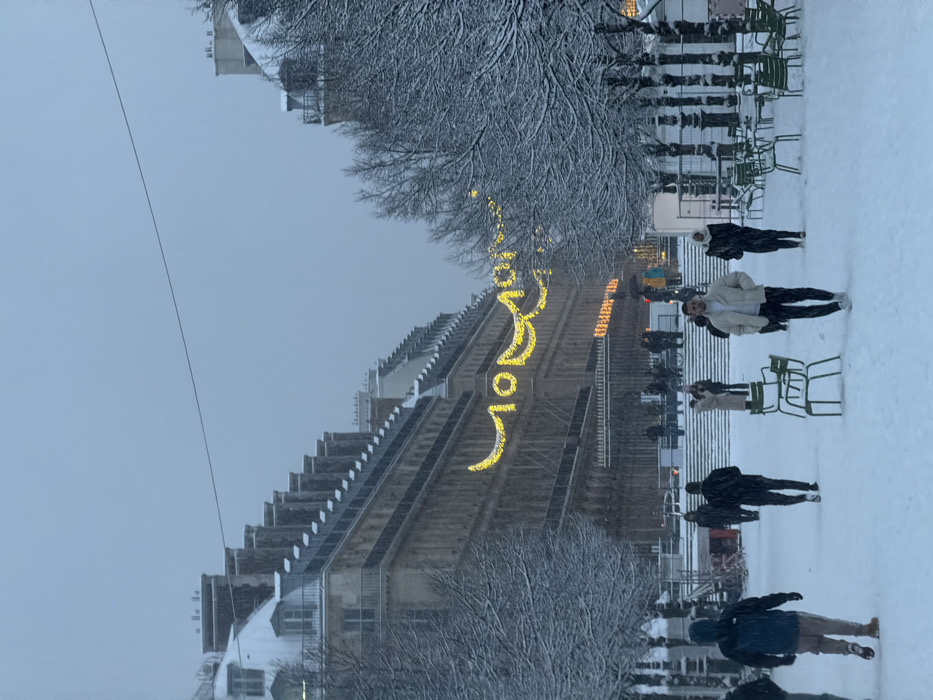 A wintery scene shows a snow-covered park with people walking along the path. Snow-dusted trees line the area. A building in the background is adorned with decorative lights. Green chairs are scattered throughout the park. The sky is overcast, contributing to the wintry atmosphere.