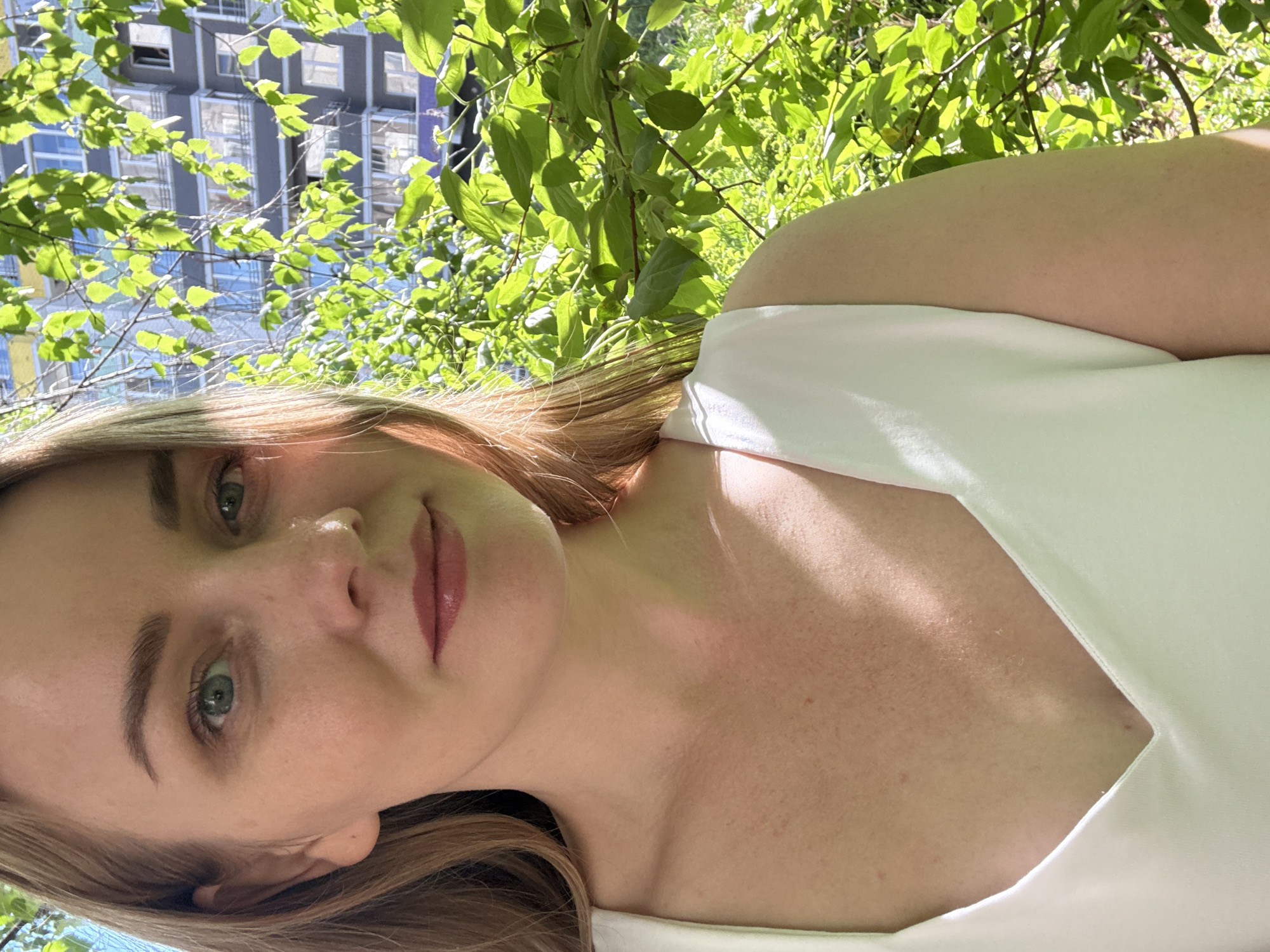 A light-skinned woman with blonde hair is posing for a selfie outdoors in the daytime. She's wearing a white garment with a V-neck cut. The background includes green foliage and a tall building with a repeating window pattern. The building and greenery could indicate an urban park or residential area in Chaikovsky, Russia. The photo composition suggests the woman is holding the camera at arm's length, taking a selfie.