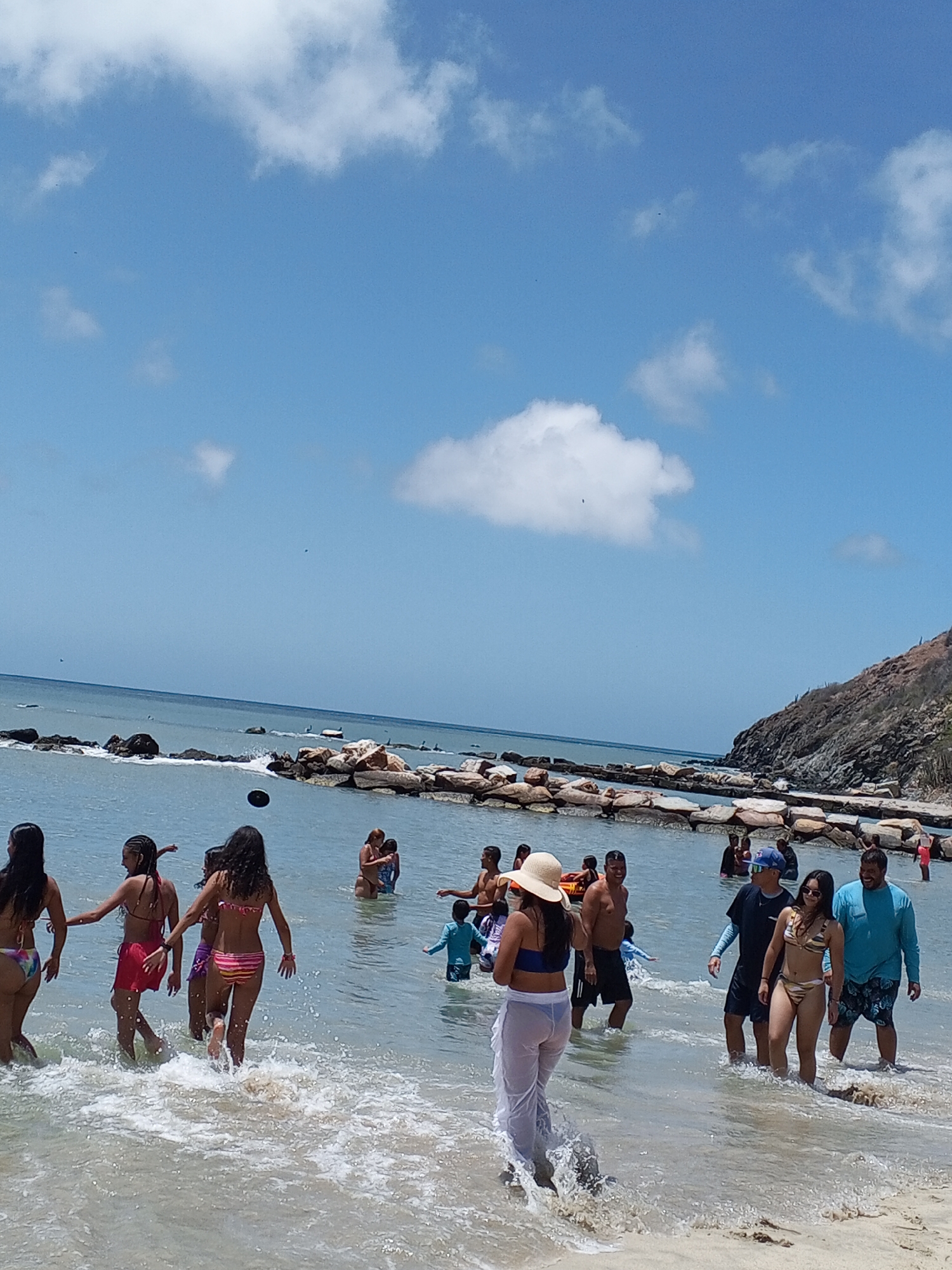 The image shows a beach scene in Pedro González, Venezuela. Numerous people of various ages and body types are in the water, which is clear and shallow near the shore. Some are wading, others are standing, and a few are actively playing or interacting. Most are wearing swimwear.
There is a group of women in the foreground, seemingly walking into the water. One woman is wearing a wide-brimmed hat and a sheer white cover-up over her bikini. Further in the background, there is a man in a black shirt and another in a light blue shirt.
The shoreline is defined by rocks and a low hill covered with sparse vegetation. The sky is blue with scattered clouds, creating a sunny day. The water extends to the horizon, where it meets the sky.